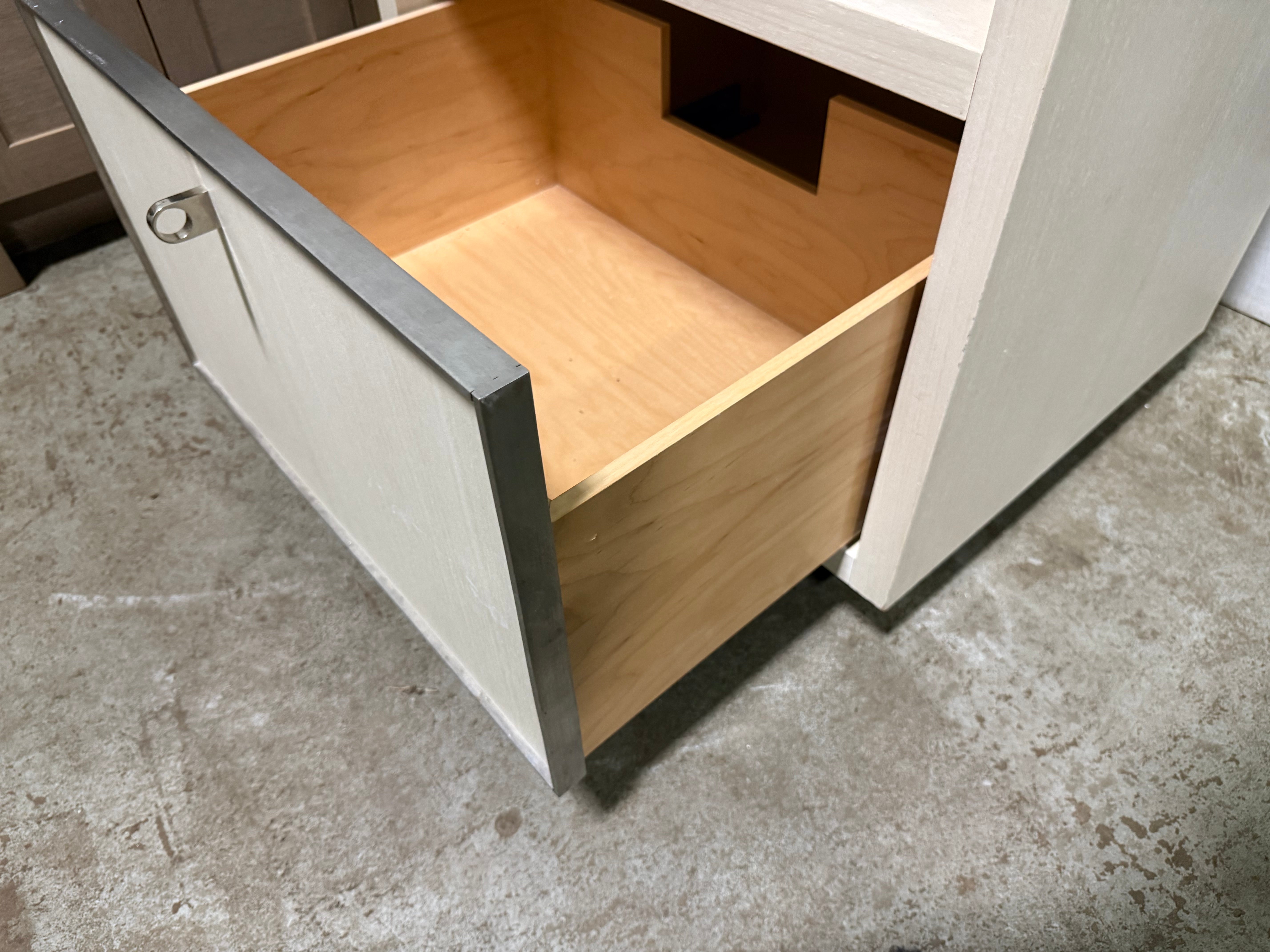A close-up of a light wood drawer with a cream-colored exterior and a dark gray metal frame, shown with the drawer pulled open on a concrete floor.