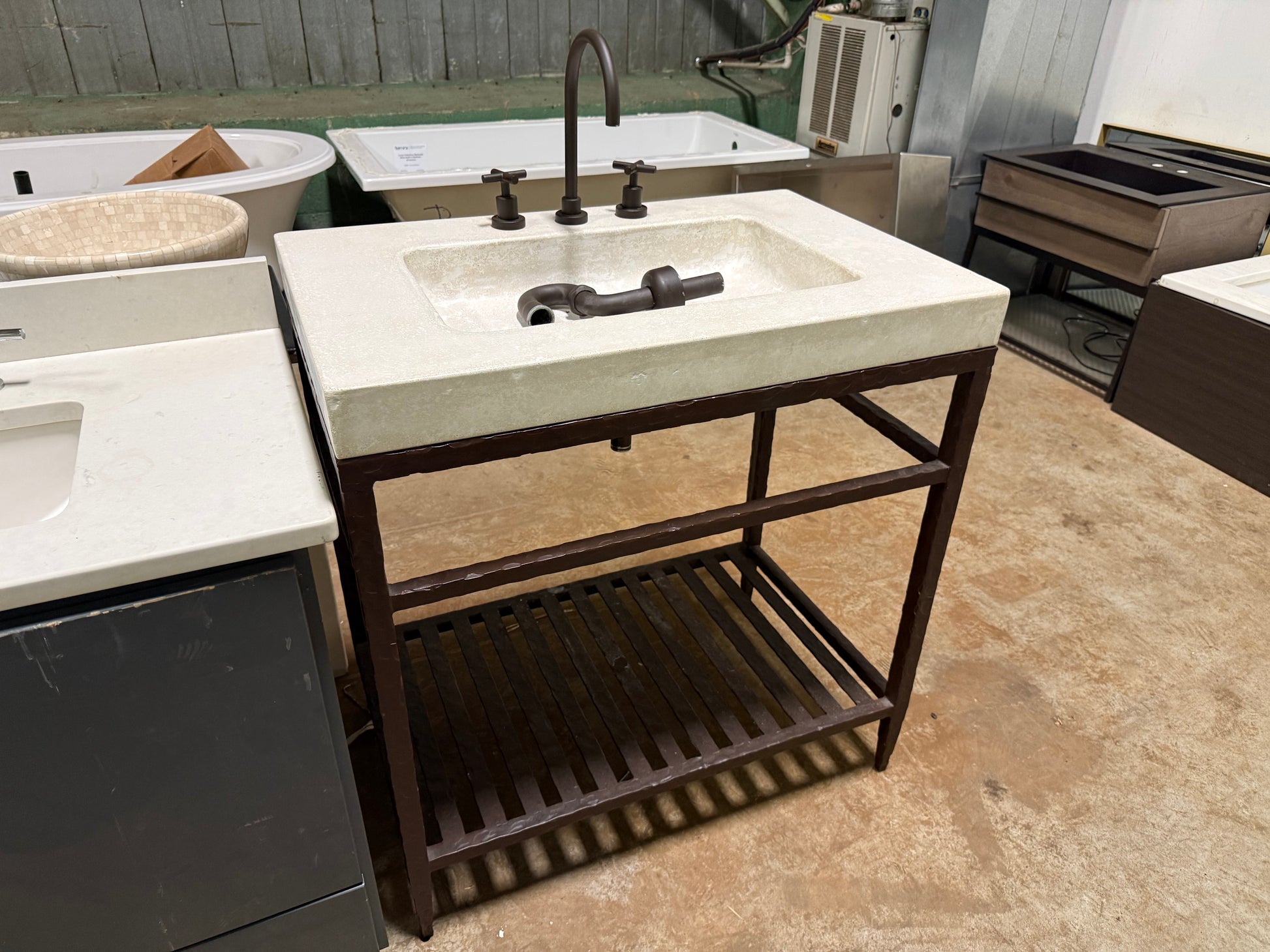A freestanding rectangular concrete-look vanity top with an integrated sink and dark metal faucet, mounted on a dark wood or metal frame with an open lower shelf, photographed in a showroom or warehouse setting.