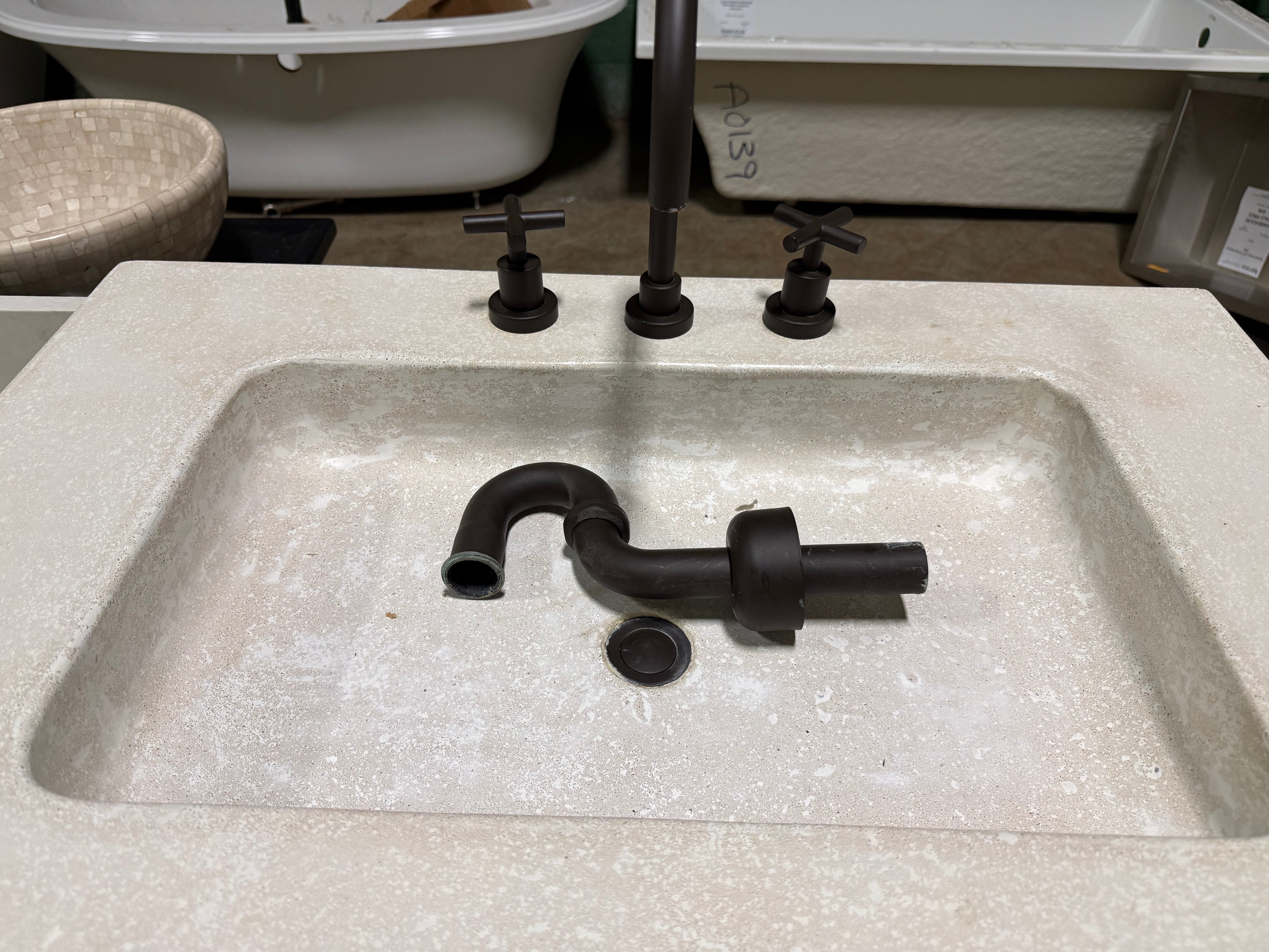 A dark bronze faucet set with a tall central spout, two cross-handle knobs, and a curved pipe section resting inside a beige stone sink basin.