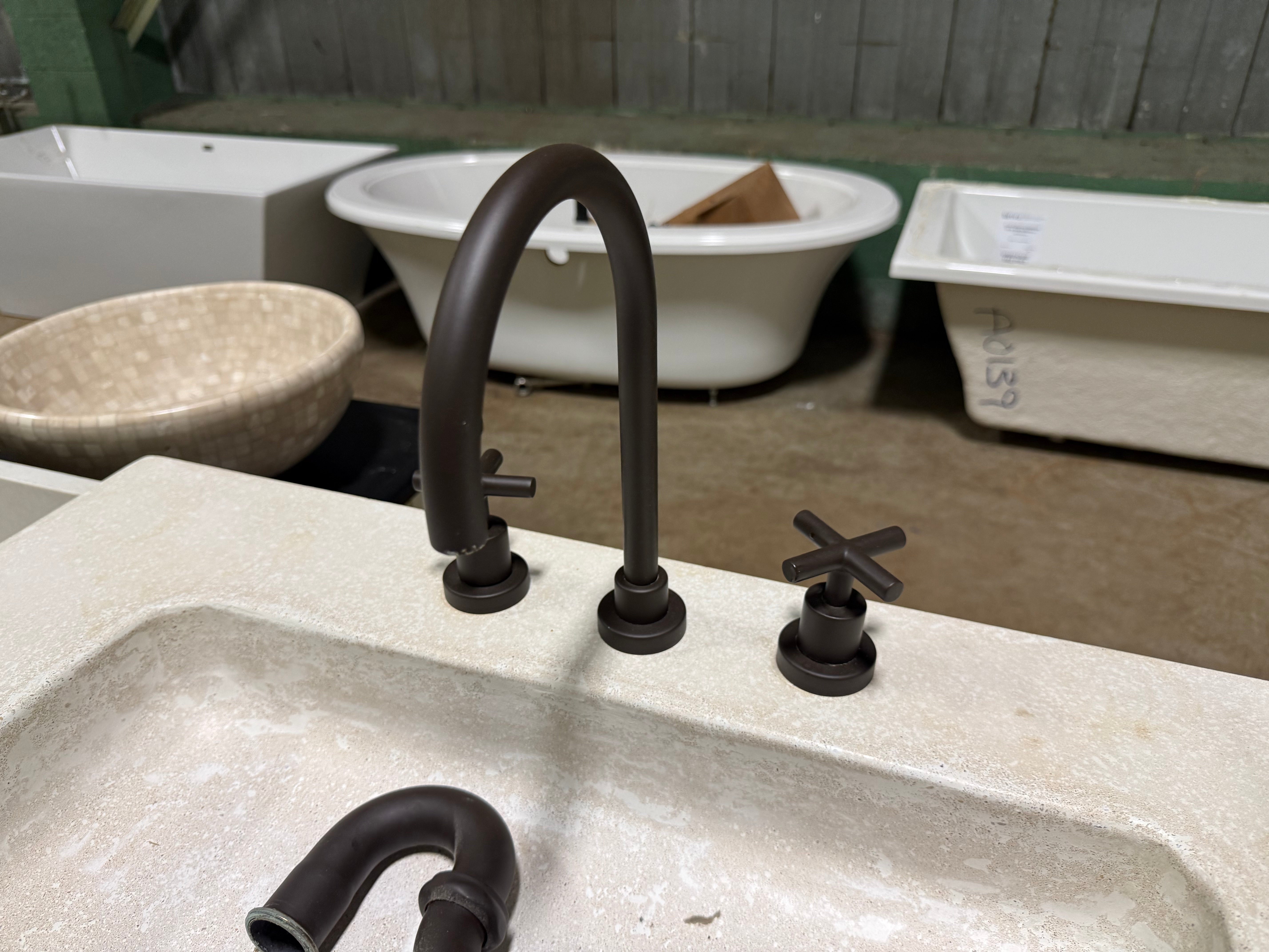 A dark bronze double-handle faucet set installed in a light beige stone sink, with a matching curved drain visible in the foreground and other bathroom fixtures in the background.