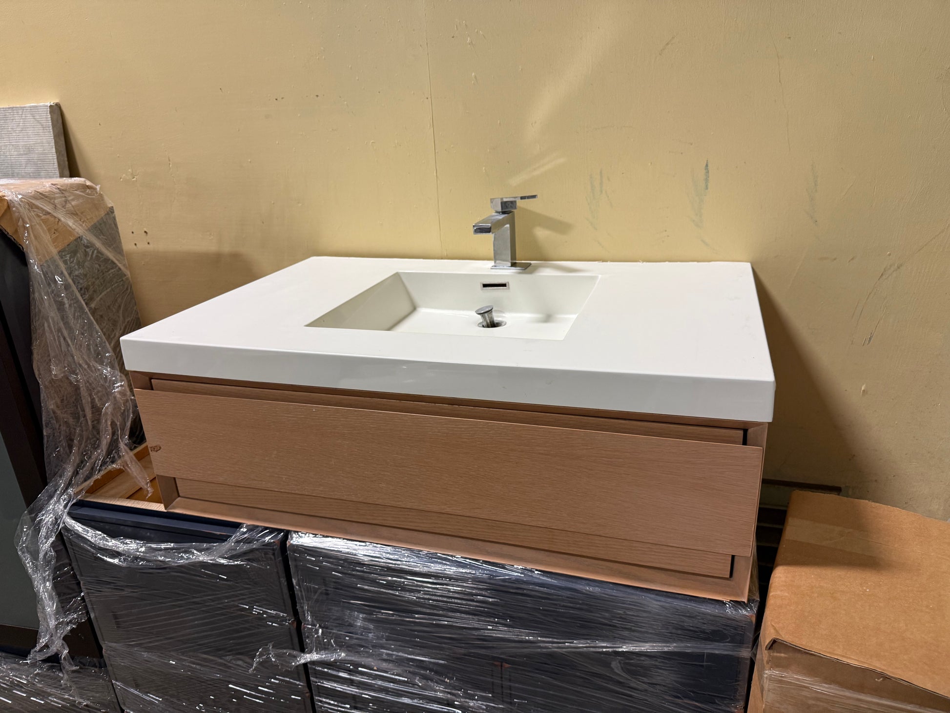 A modern bathroom vanity set with a white rectangular sink and countertop, a silver faucet, and a light wood cabinet, shown in a warehouse-like setting with protective plastic wrapping on parts of the furniture.