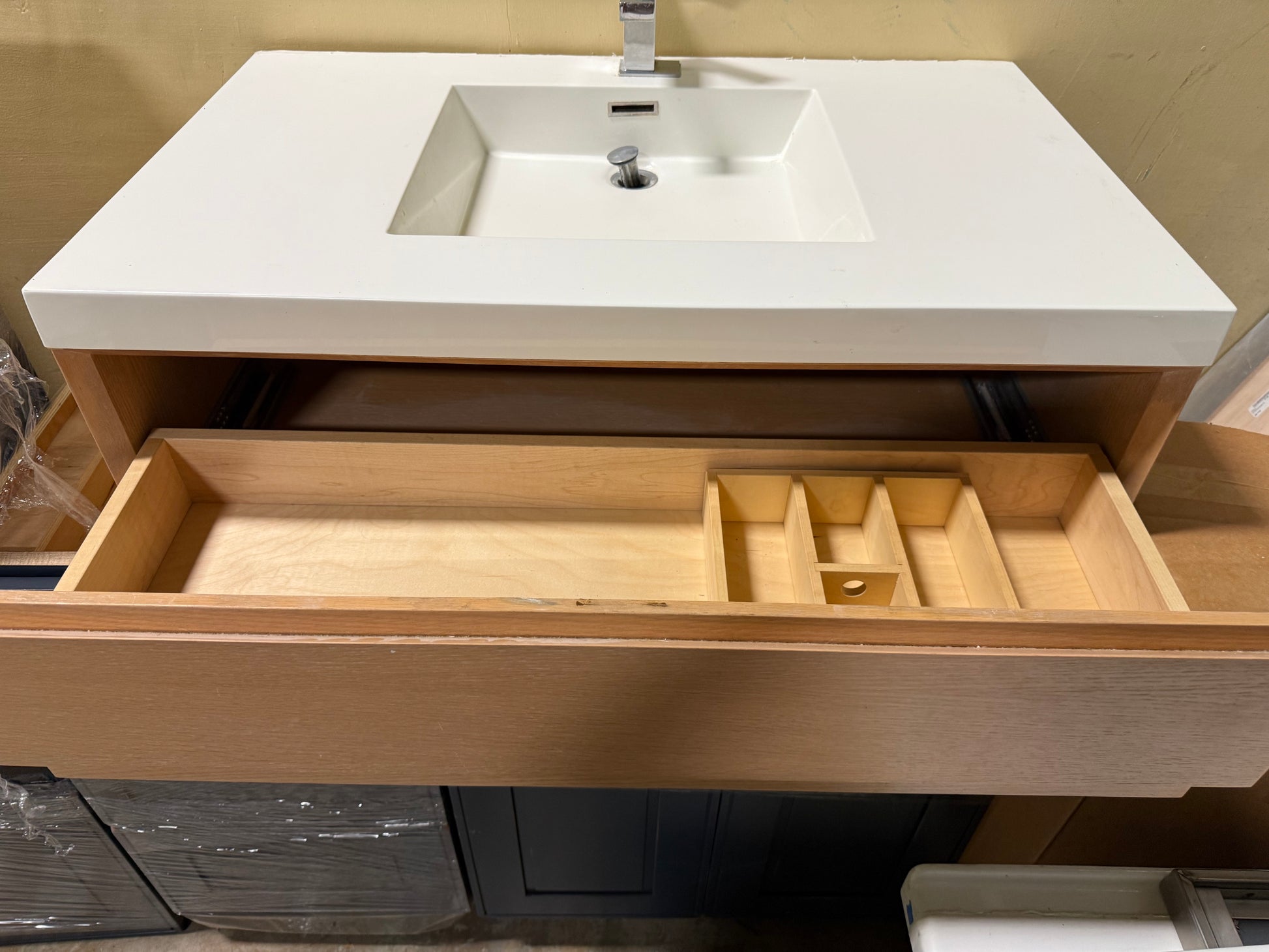 A modern bathroom vanity with a white rectangular sink and countertop, a silver faucet, and an open light wood drawer showing a divider tray inside.