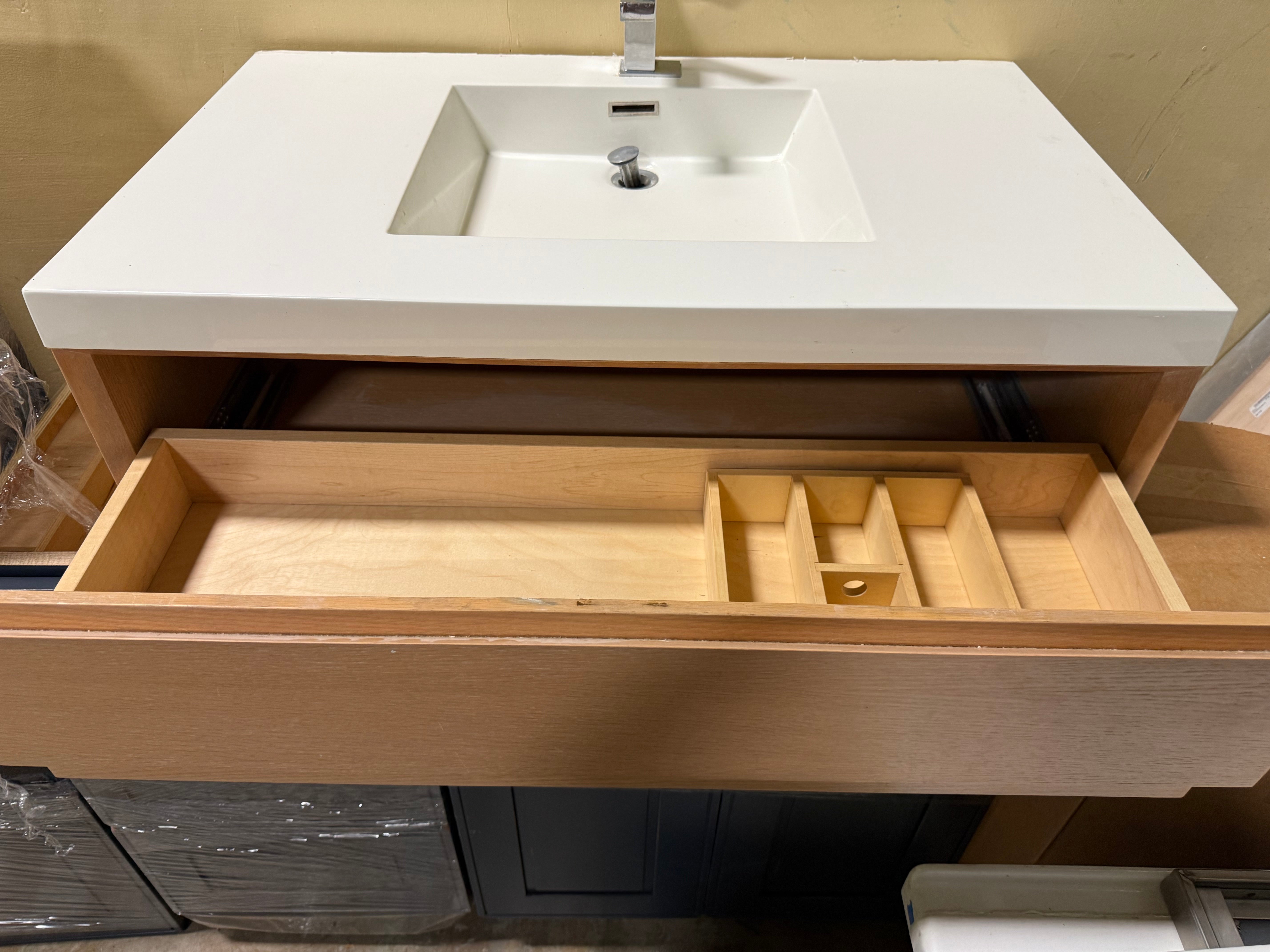 A modern bathroom vanity with a white rectangular sink and countertop, a silver faucet, and an open light wood drawer showing a divider tray inside.