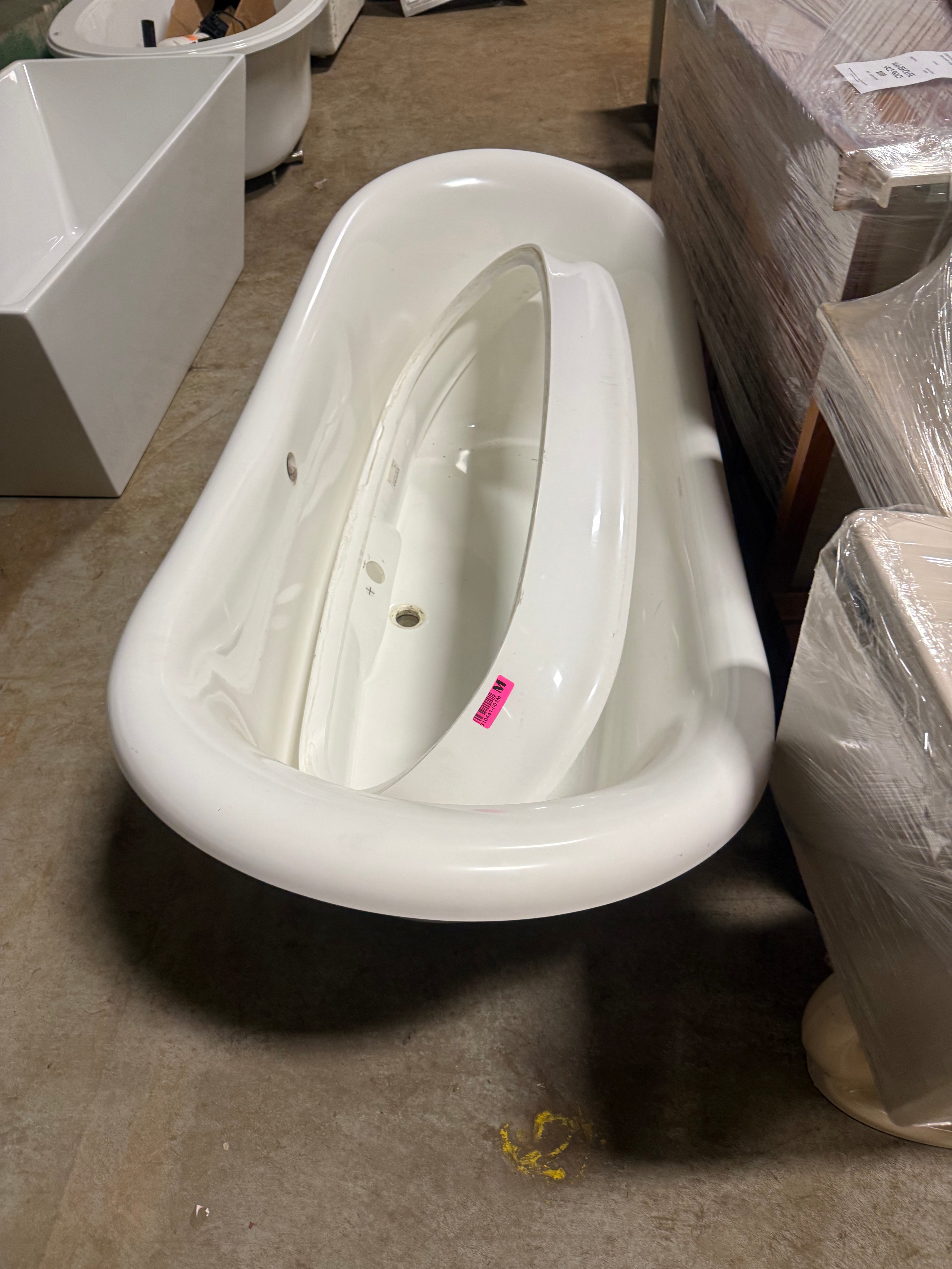 A white freestanding bathtub shown in a warehouse or showroom setting, surrounded by other tubs and wrapped furniture. A small pink sticker is visible on the interior wall of the tub.