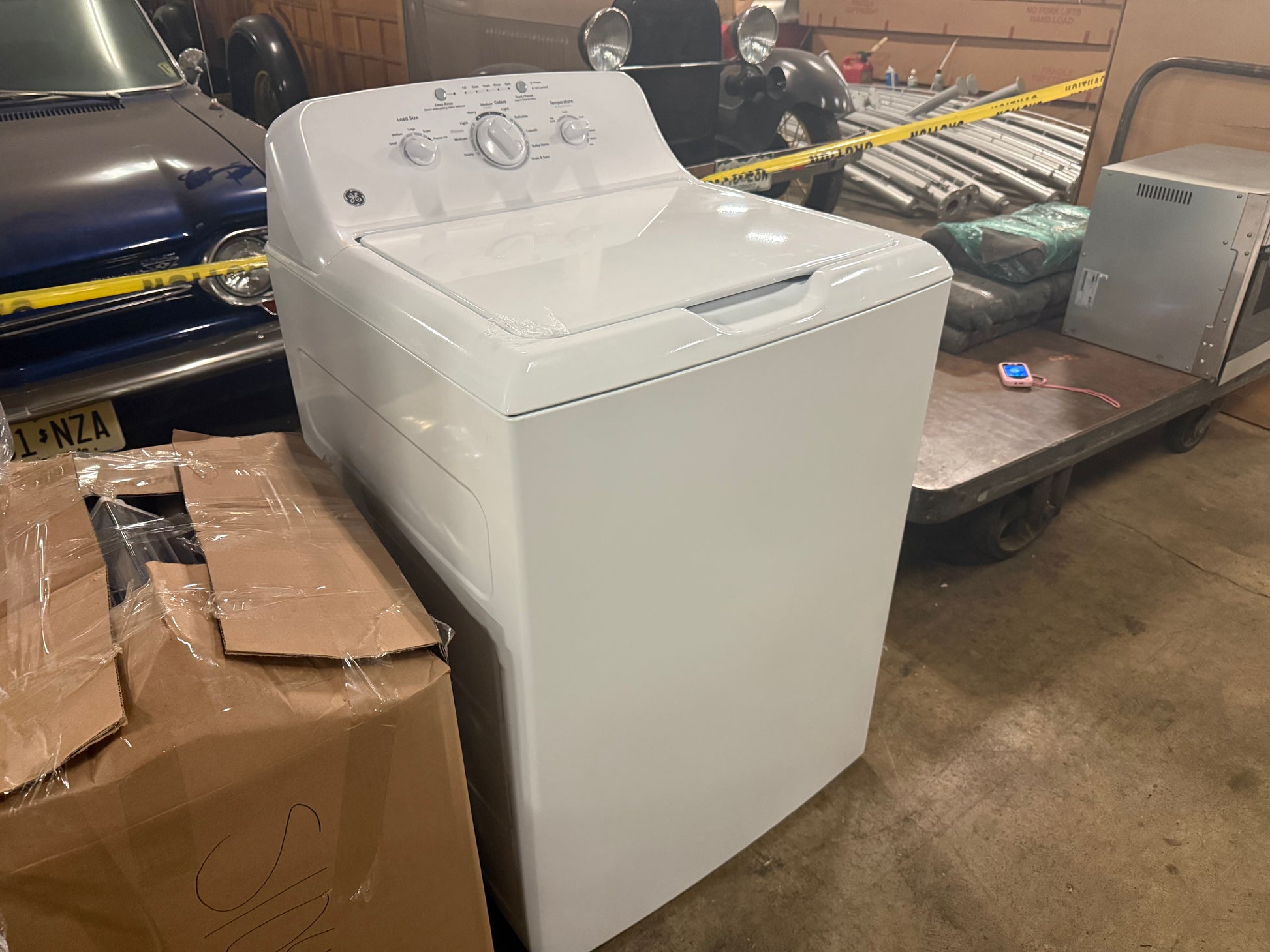 A white top-loading washing machine photographed in a warehouse or garage setting, with cardboard boxes, a blue car, and a cart visible around it.