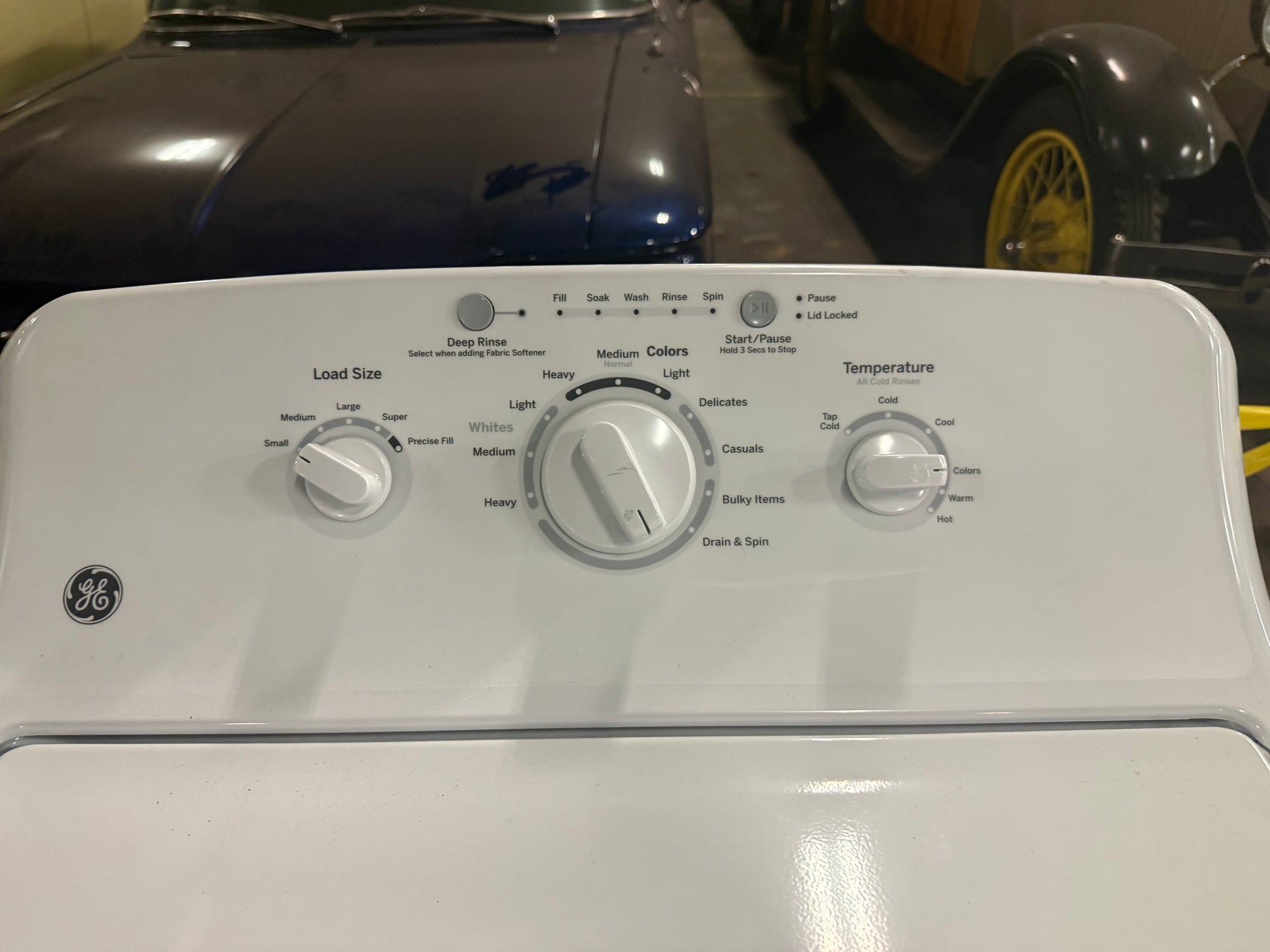 A white GE top-loading washing machine shown from the front, with control knobs and labels visible on the console. The background is a garage setting with a dark car and another vehicle wheel visible behind it.