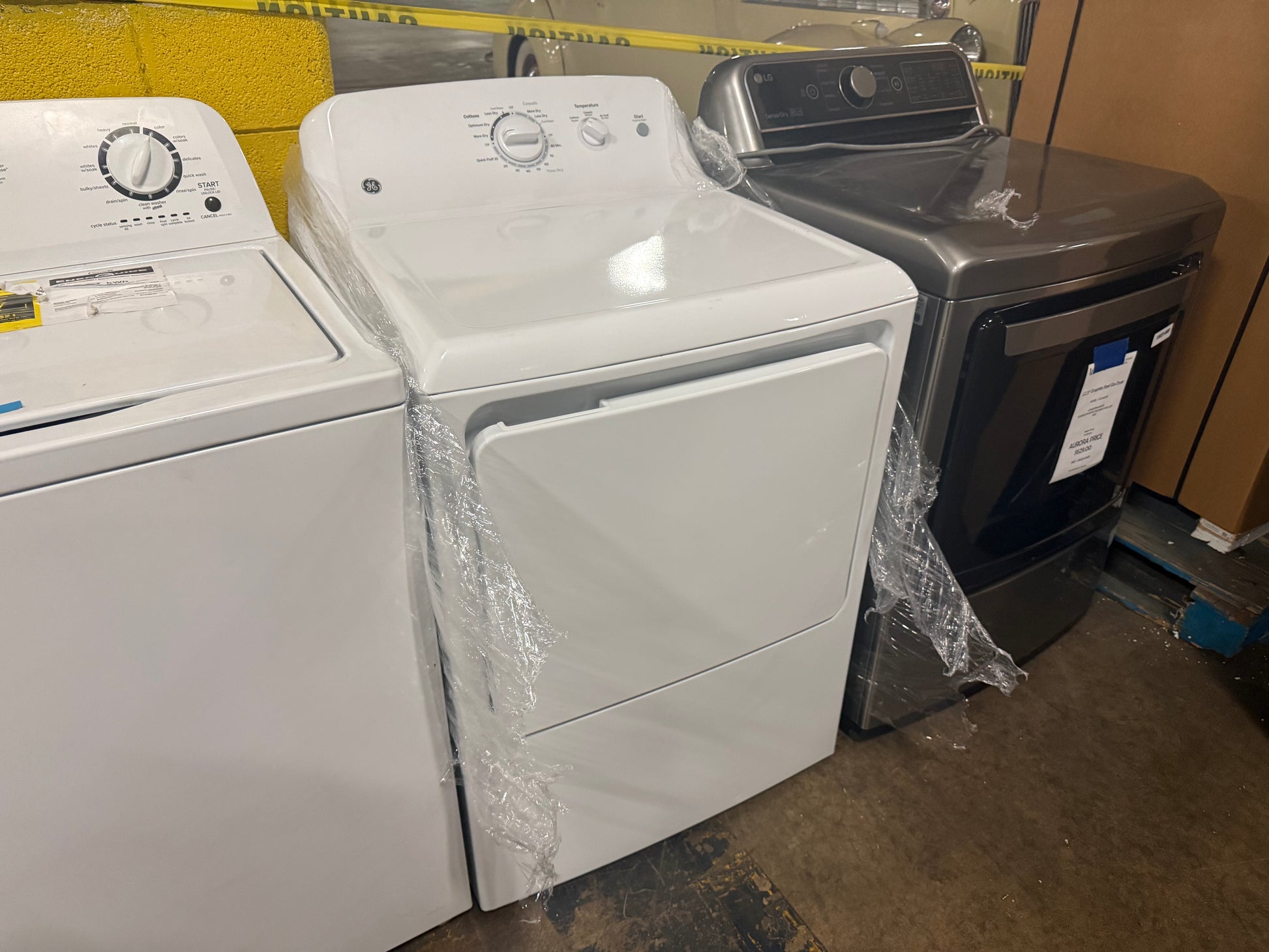 A white top-loading washing machine and a matching white front-loading dryer are shown side by side, both still wrapped in protective plastic. A dark gray/black clothes dryer is partially visible behind them in what appears to be a retail or storage area.