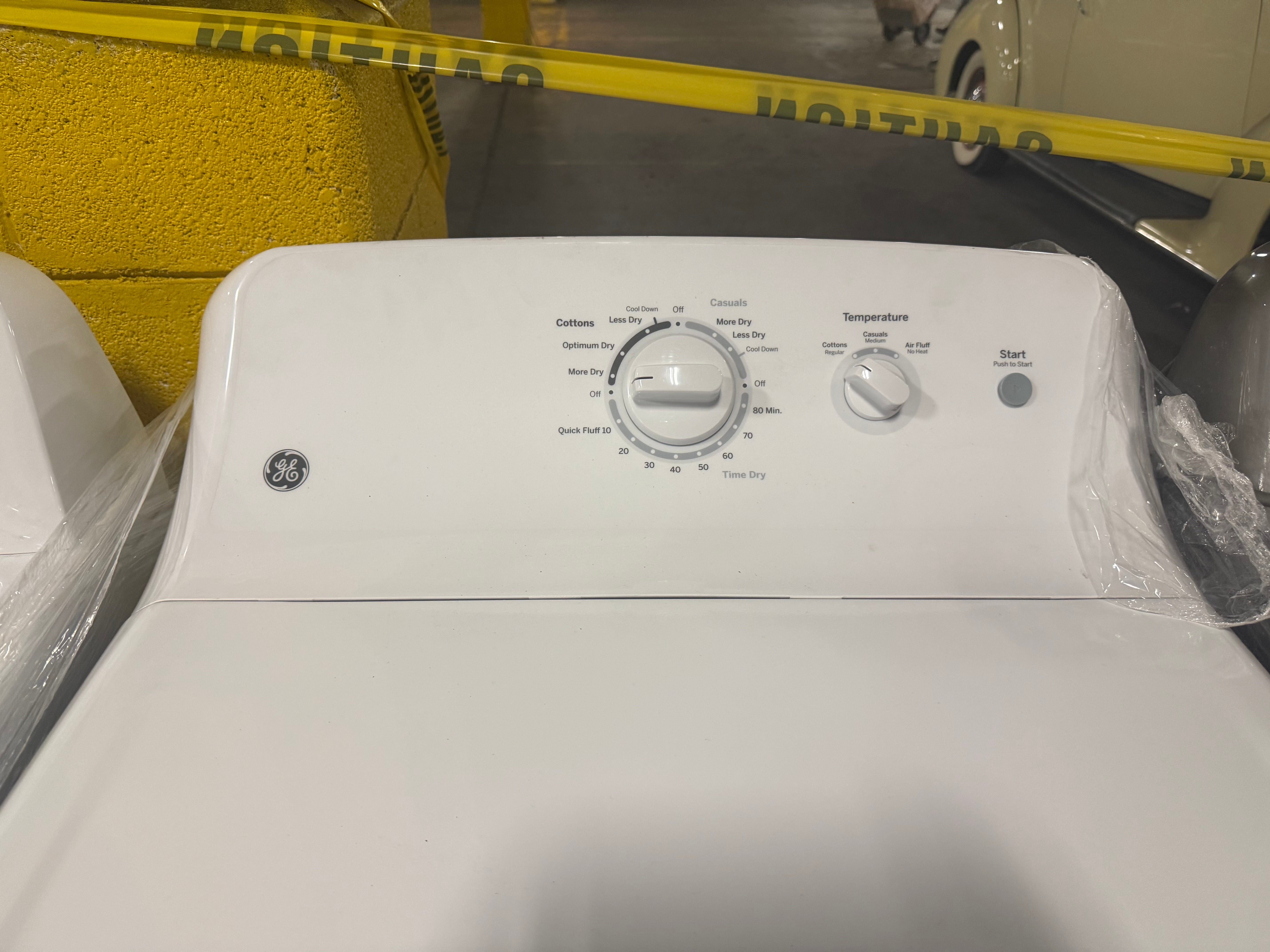A white GE top-loading washing machine photographed indoors, with the control panel facing forward and a dial, temperature setting, and start button visible. The appliance is partially surrounded by protective plastic, with a yellow wall and store interior in the background.