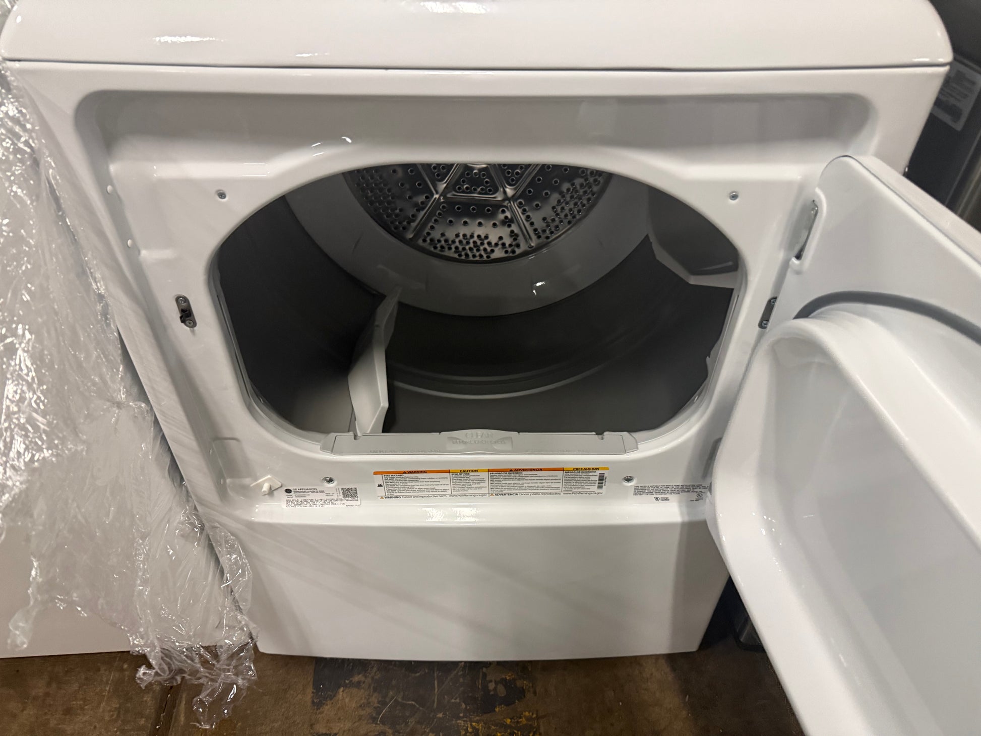 A white front-loading clothes dryer with its door open, showing the interior drum and lint trap area. The dryer is photographed indoors with plastic wrapping still visible on the left side.