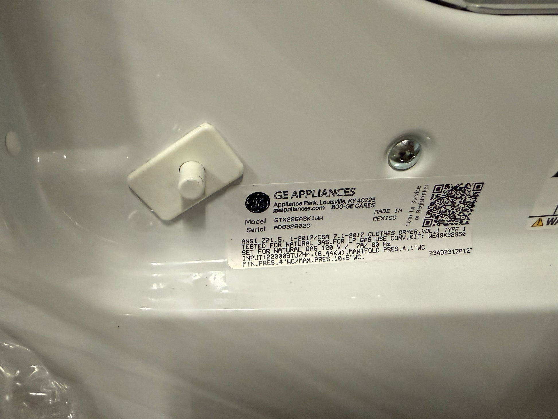 A close-up of a GE Appliances appliance label sticker on a white plastic surface, showing model information, a QR code, and a small white plastic fastener.