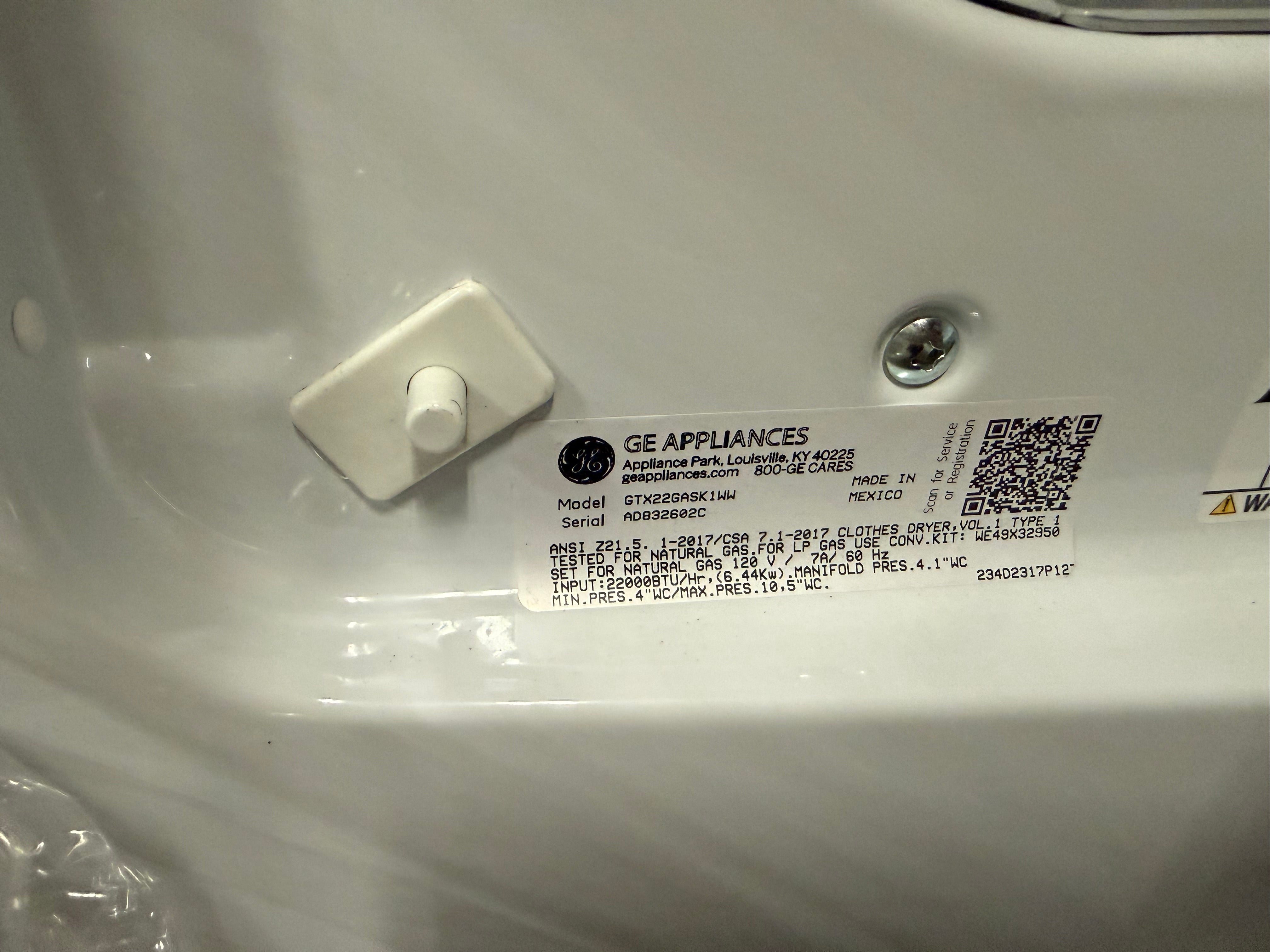 A close-up of a GE Appliances appliance label sticker on a white plastic surface, showing model information, a QR code, and a small white plastic fastener.