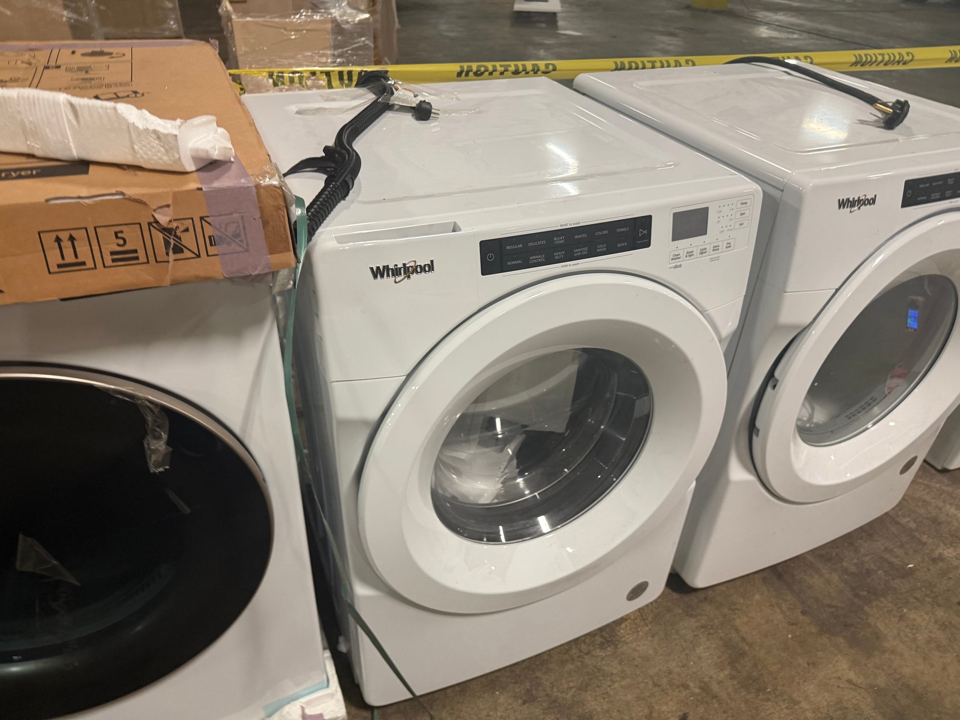A white Whirlpool front-load washing machine shown in a warehouse or showroom setting, with a cardboard box stacked on top of another appliance to the left.