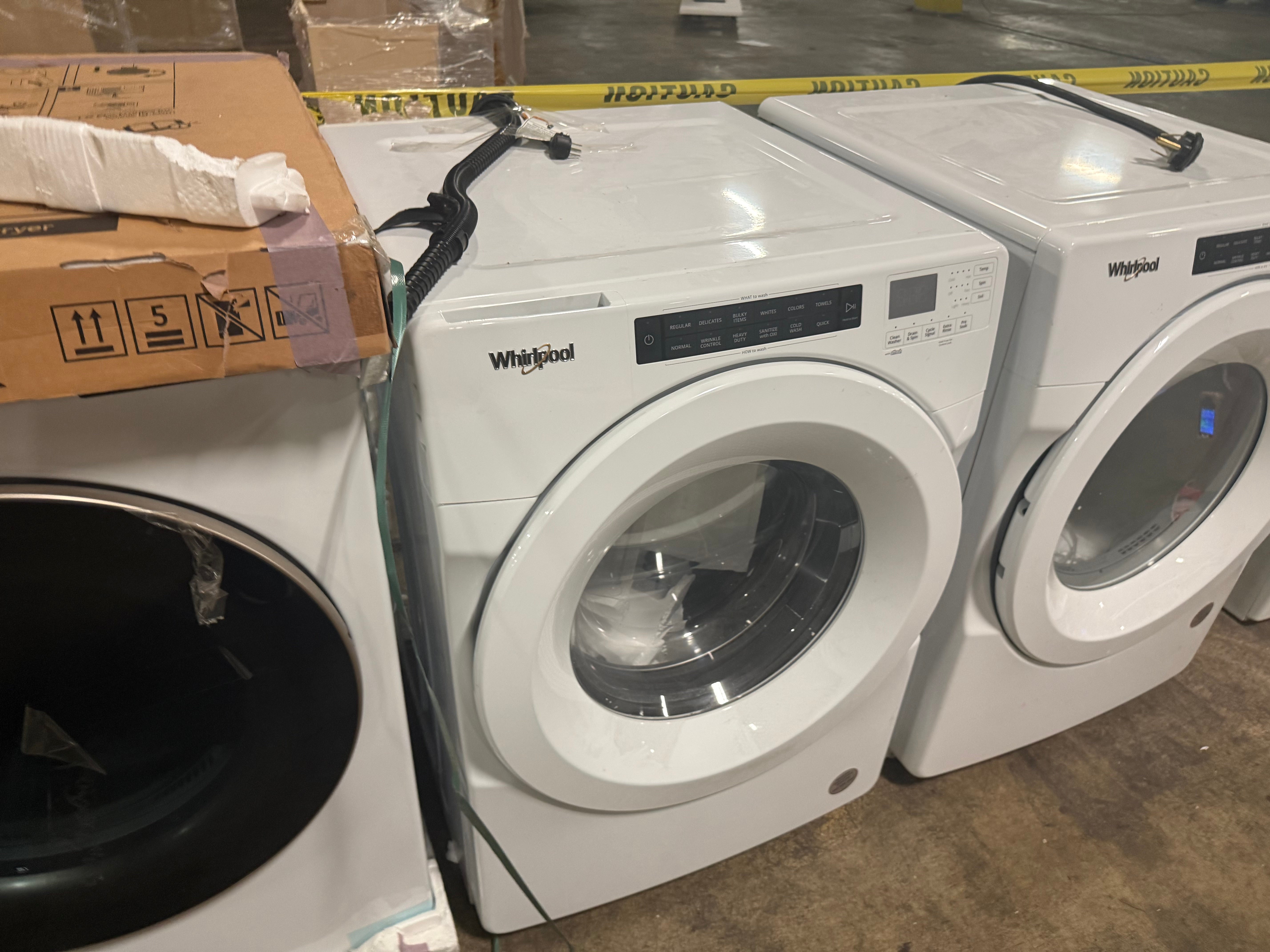 A white Whirlpool front-load washing machine shown in a warehouse or showroom setting, with a cardboard box stacked on top of another appliance to the left.