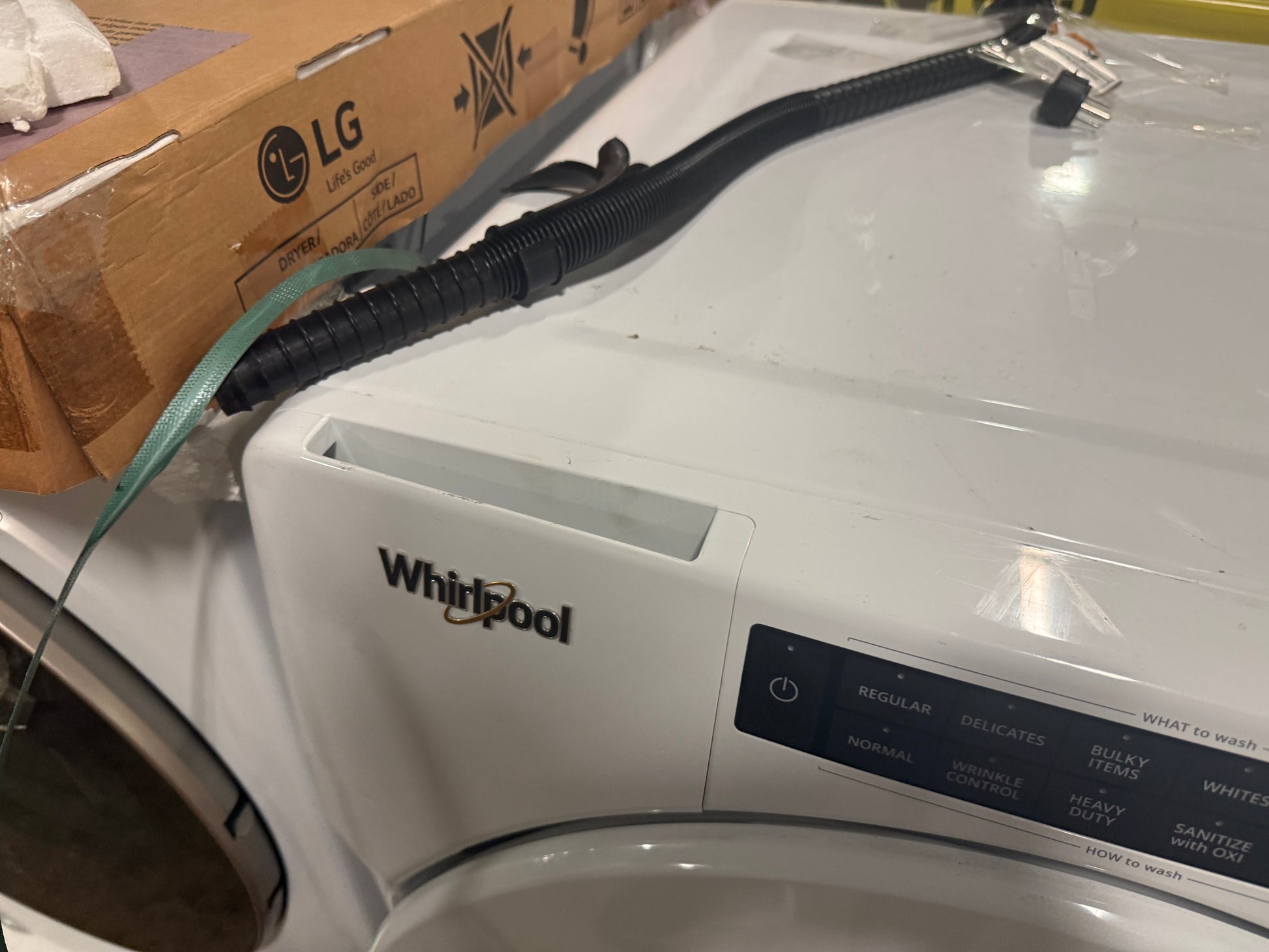 A white Whirlpool front-load washing machine with a black drain hose resting on top, next to a cardboard LG box.