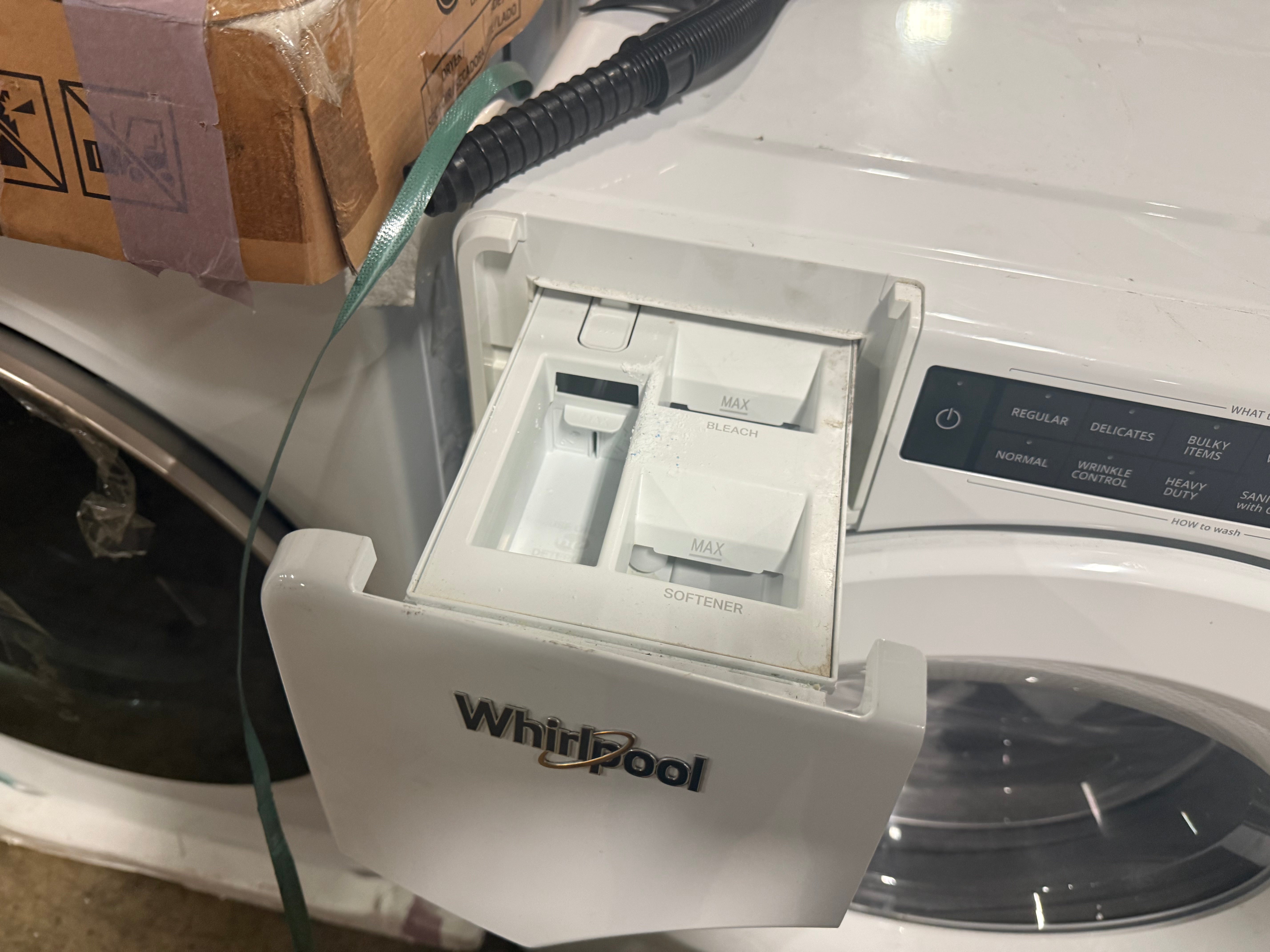 A white Whirlpool washing machine with the detergent drawer open, showing the inside compartments. A cardboard box is visible in the upper left corner, and a green cord runs down the side.