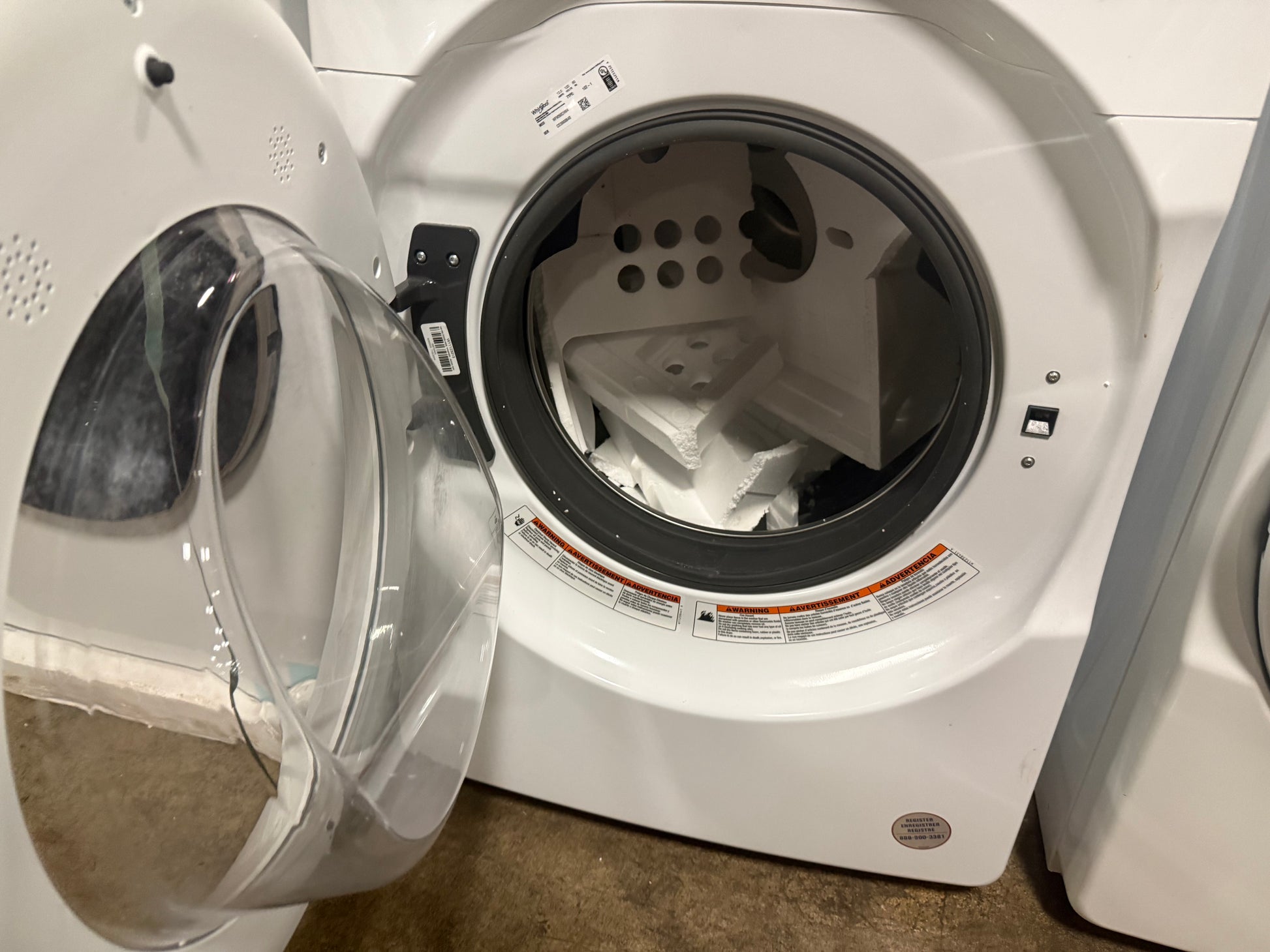 A white front-loading washing machine with its door open, showing the drum and interior drum assembly. Another washing machine is partially visible on the left, and a brown floor is at the bottom.