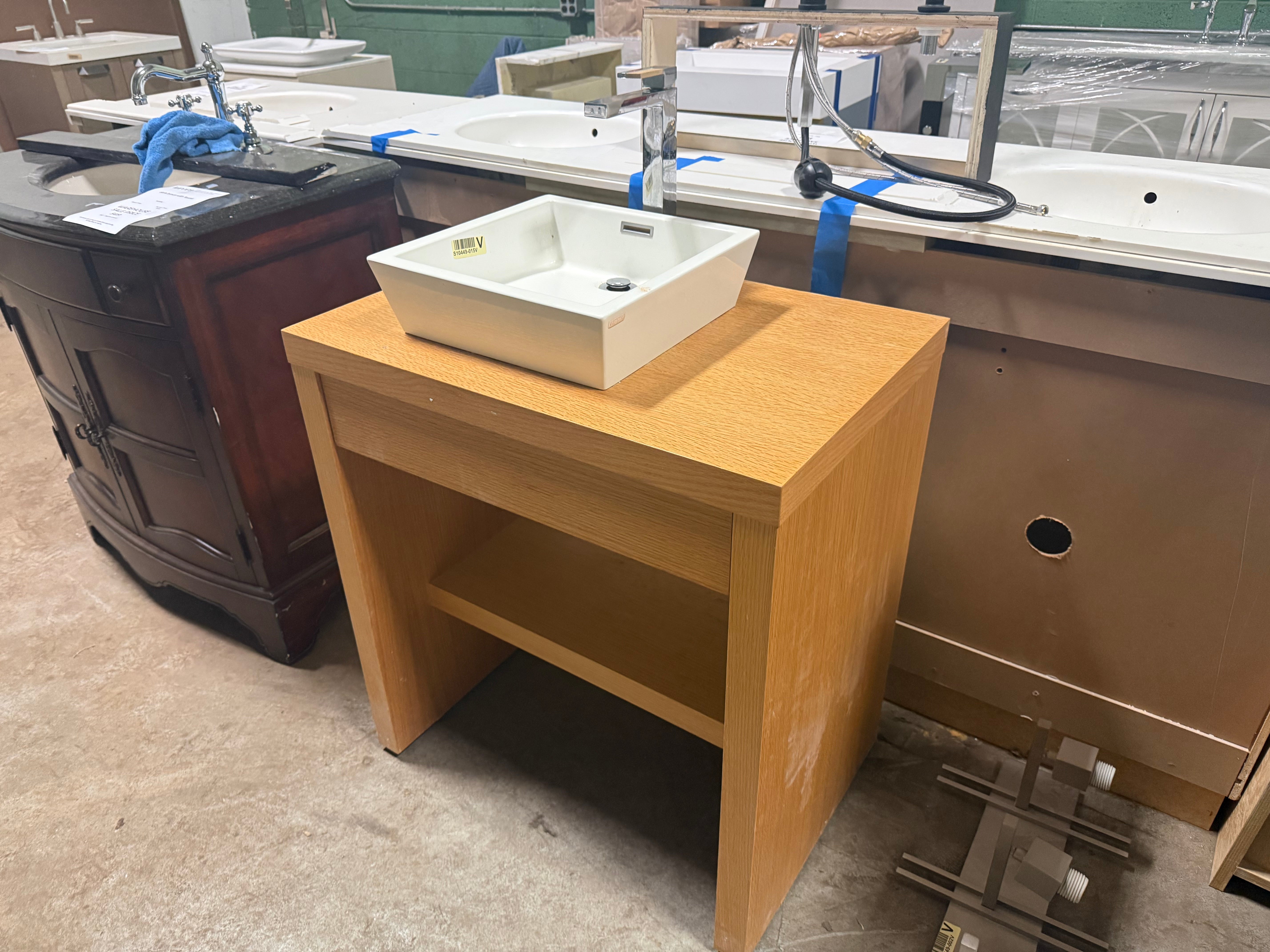 A light wood vanity cabinet with an open lower shelf, shown with a white rectangular vessel sink on top. The cabinet is photographed in a showroom or warehouse setting with other sinks and fixtures visible in the background.