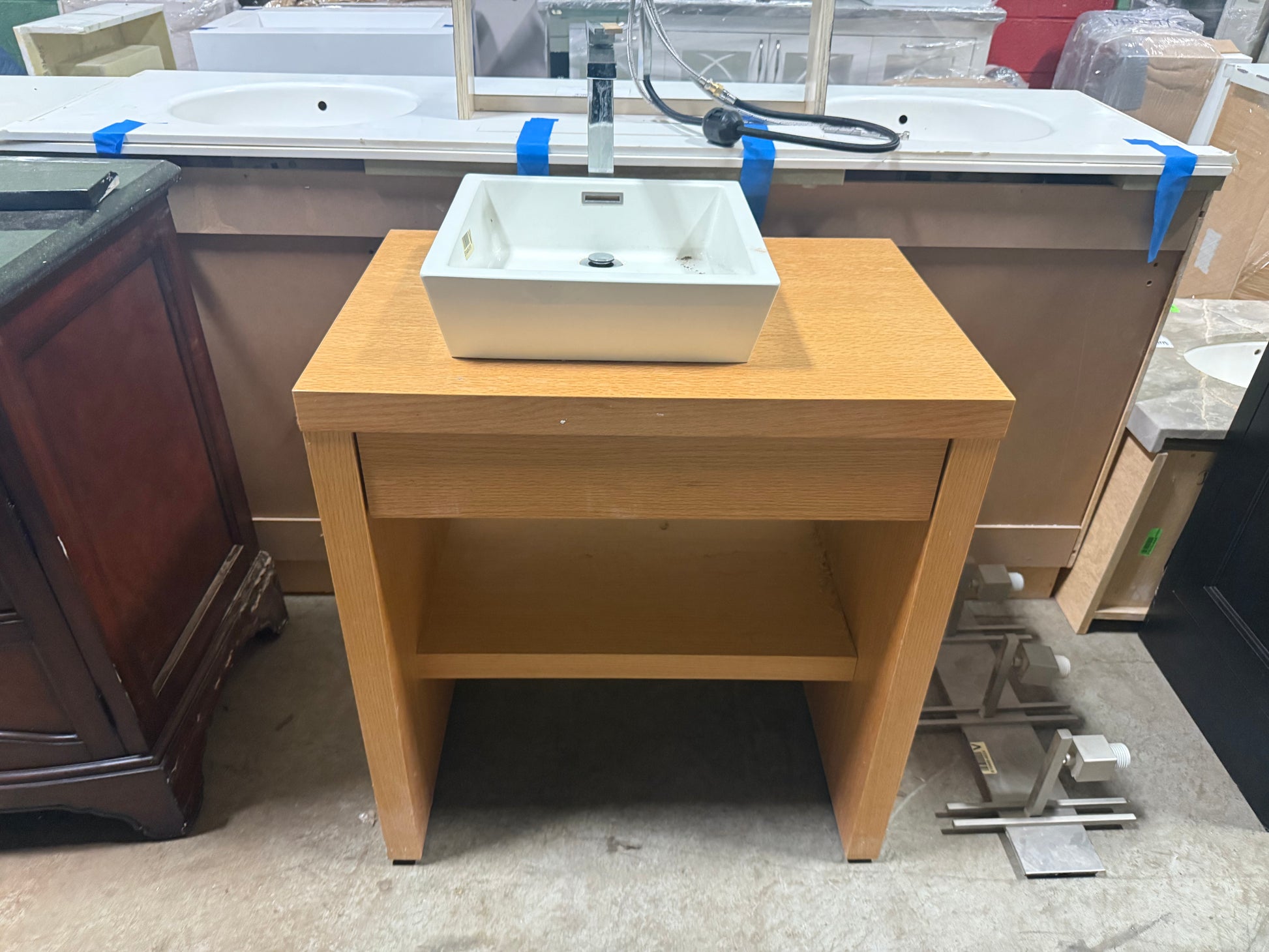 A light oak wood single-slab vanity cabinet with an open lower shelf, holding a white rectangular vessel sink and a chrome faucet, photographed in a showroom or warehouse setting.