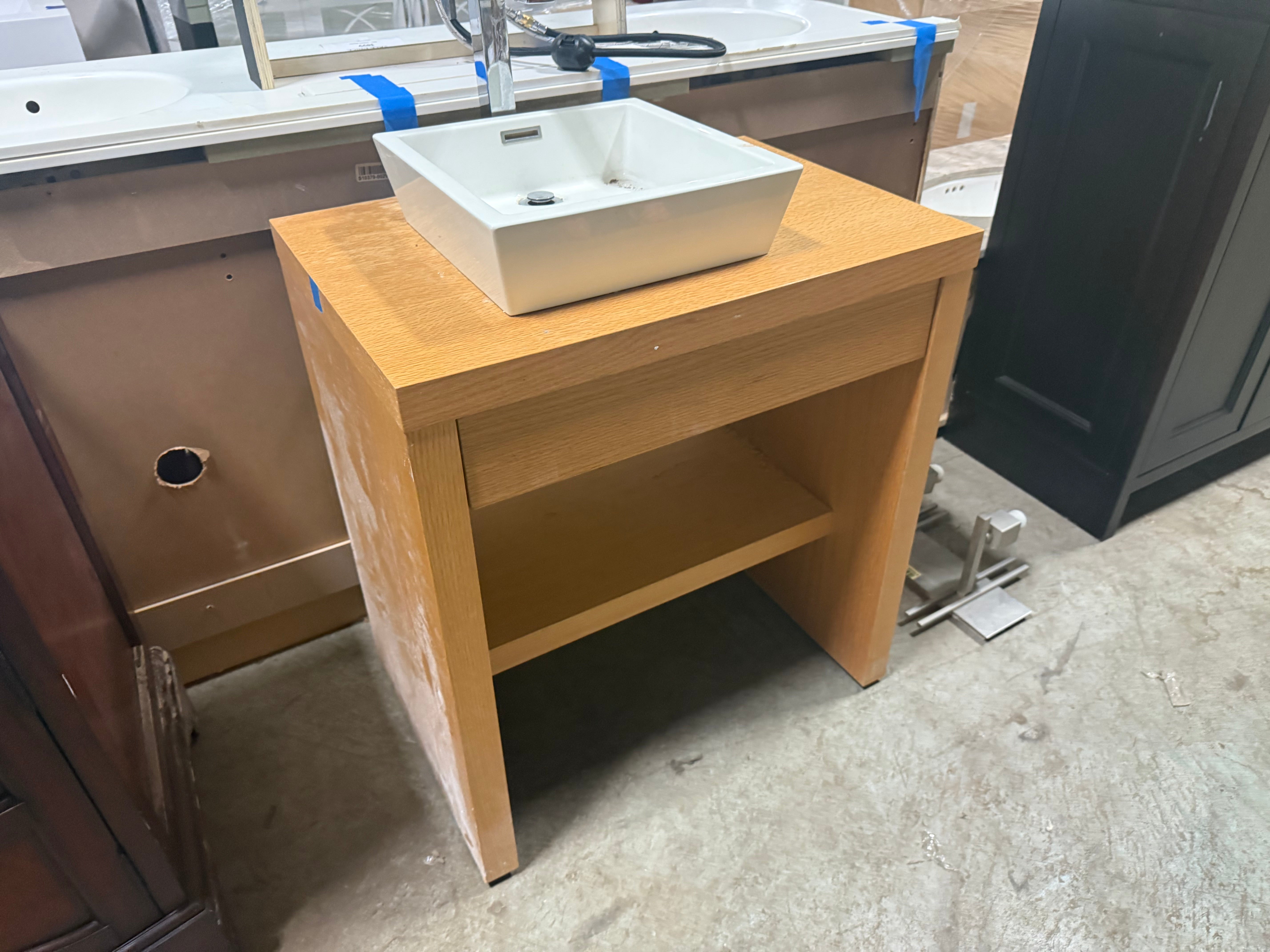 A light natural wood bathroom vanity cabinet with an open lower shelf, topped with a white rectangular vessel sink and a chrome faucet, photographed in a showroom or warehouse setting.