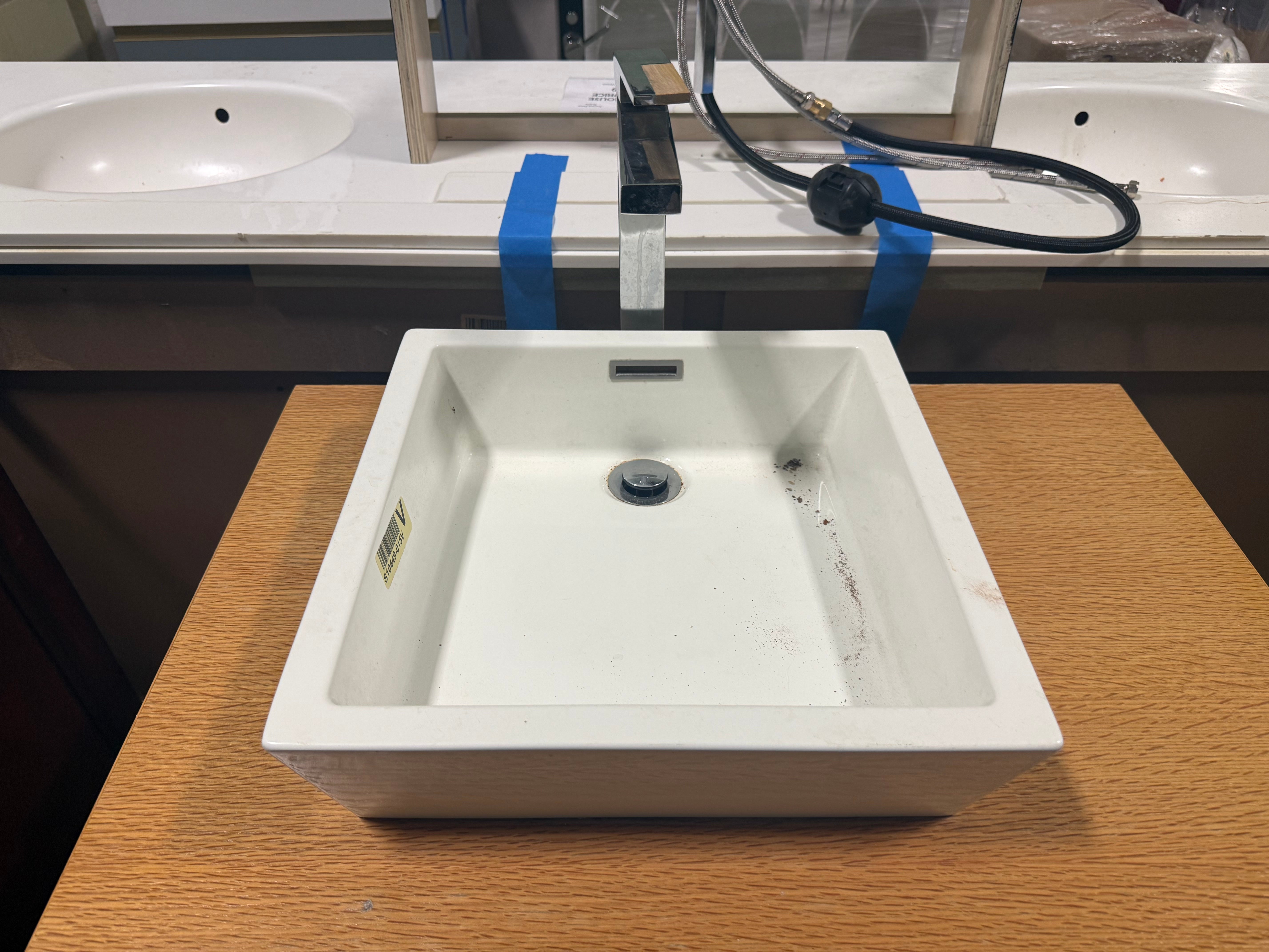 A square white ceramic bathroom sink basin displayed on a wooden table, with a faucet and plumbing cords visible behind it.