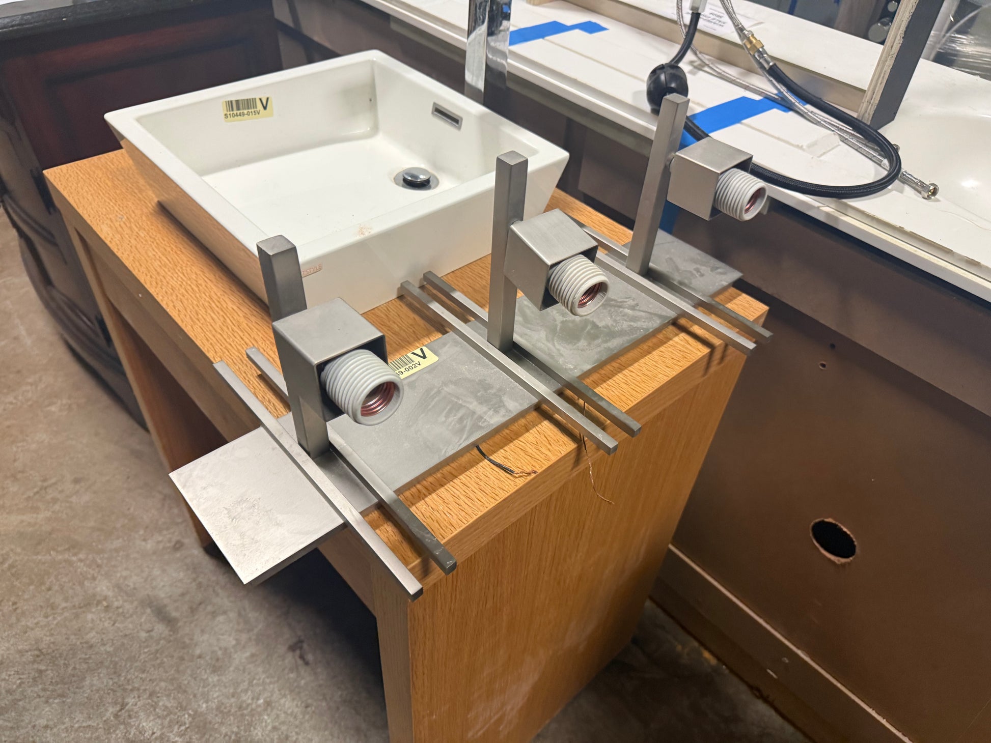 A white rectangular vessel sink basin mounted on a wooden workbench, with metal fixture brackets and a faucet hardware component attached in a workshop or showroom setting.
