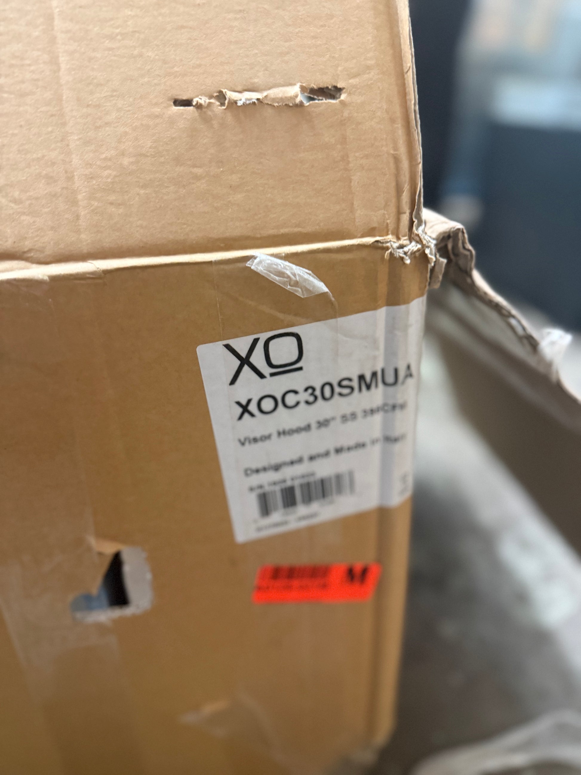 A close-up of a brown cardboard shipping box with a white label reading 'XQ XOC30SMUA' and a small orange shipping sticker near the bottom.