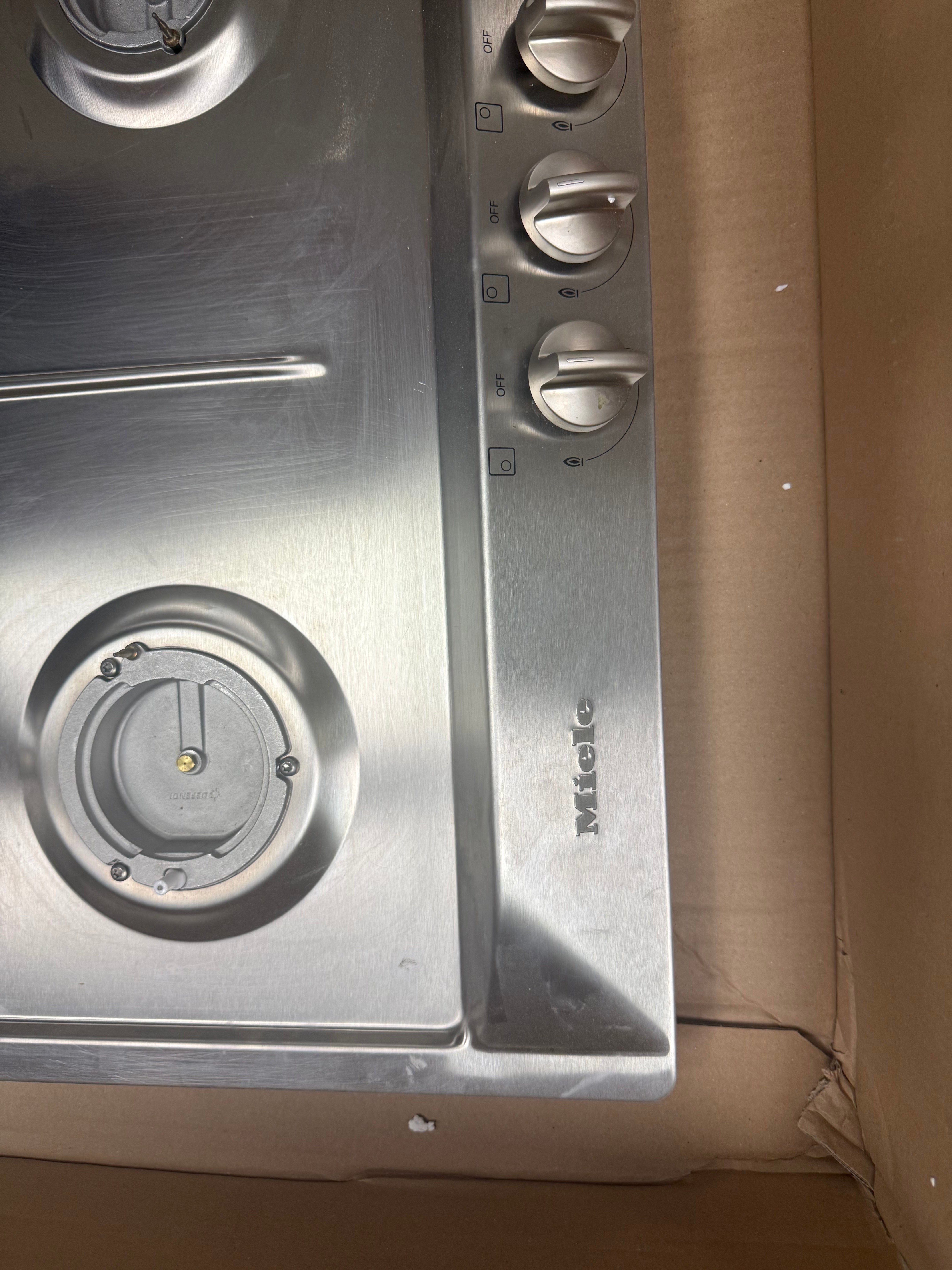 A close-up of a stainless steel gas cooktop or stove burner area with three control knobs on the right side and a visible Miele logo, resting on a brown cardboard box.