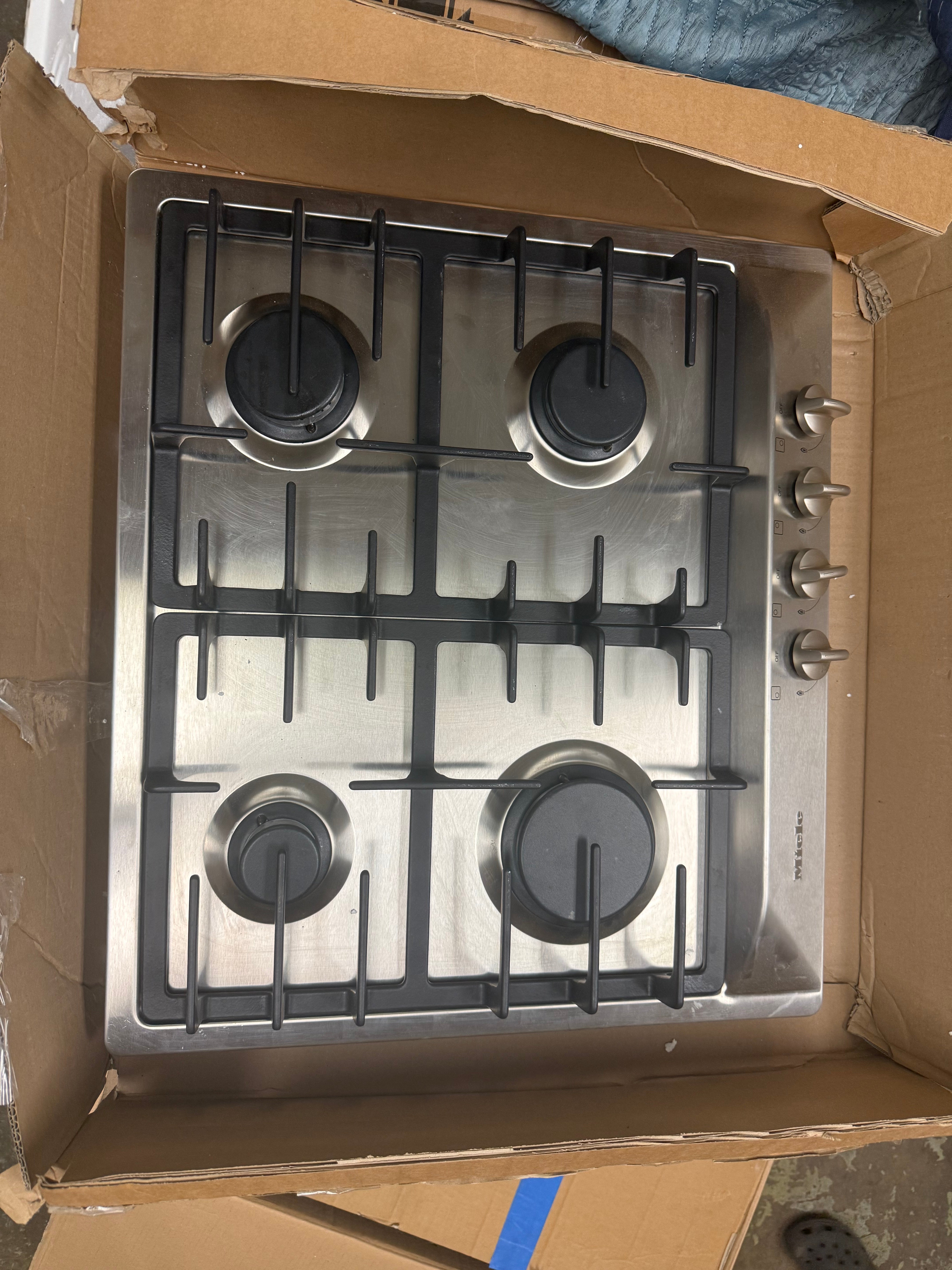 A stainless steel 4-burner gas cooktop with black grates and control knobs, shown inside an open cardboard box.
