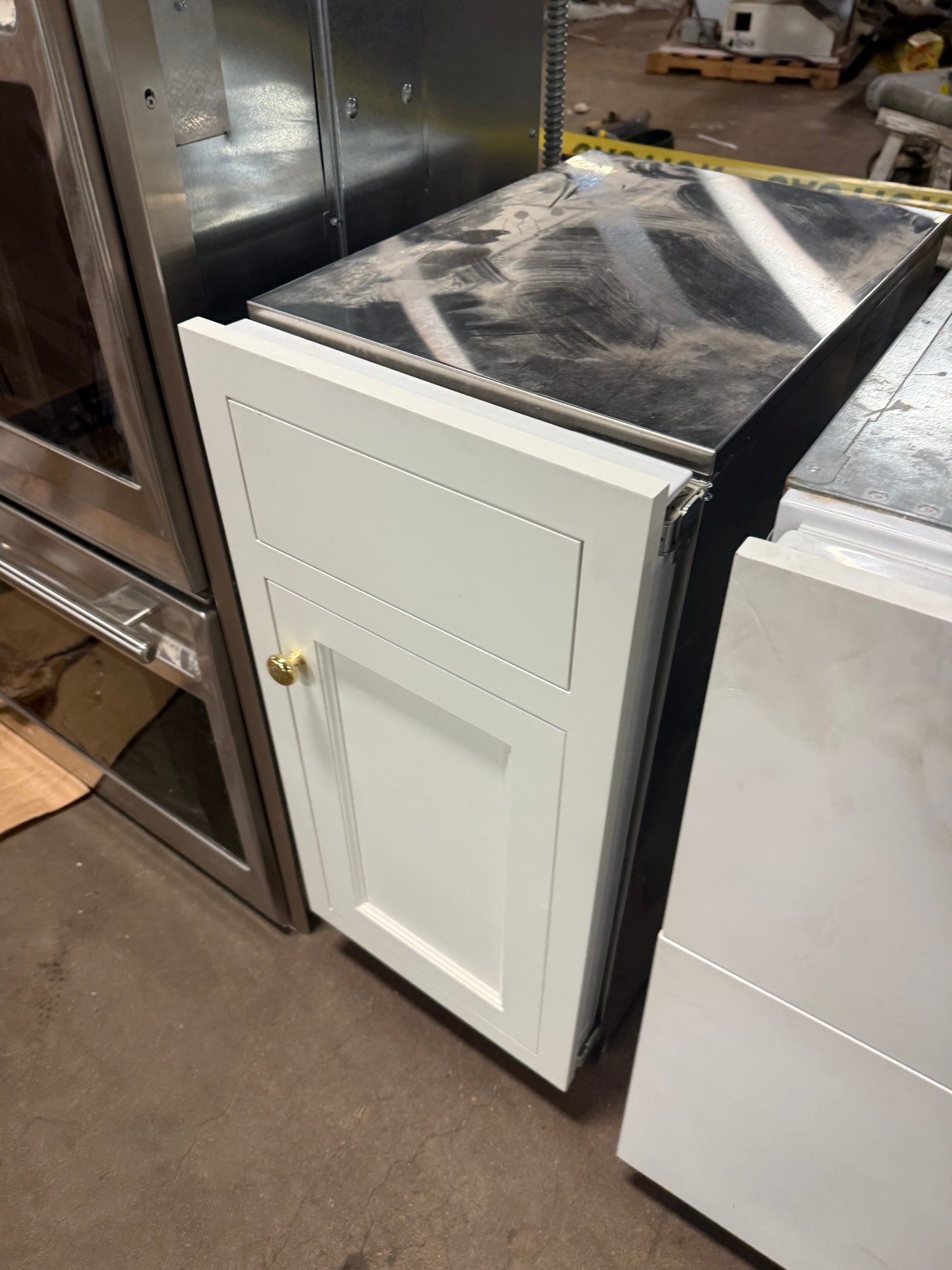 A white single cabinet base with a recessed panel door and a small brass knob, shown installed or staged next to other cabinetry and appliances in a warehouse-like setting.