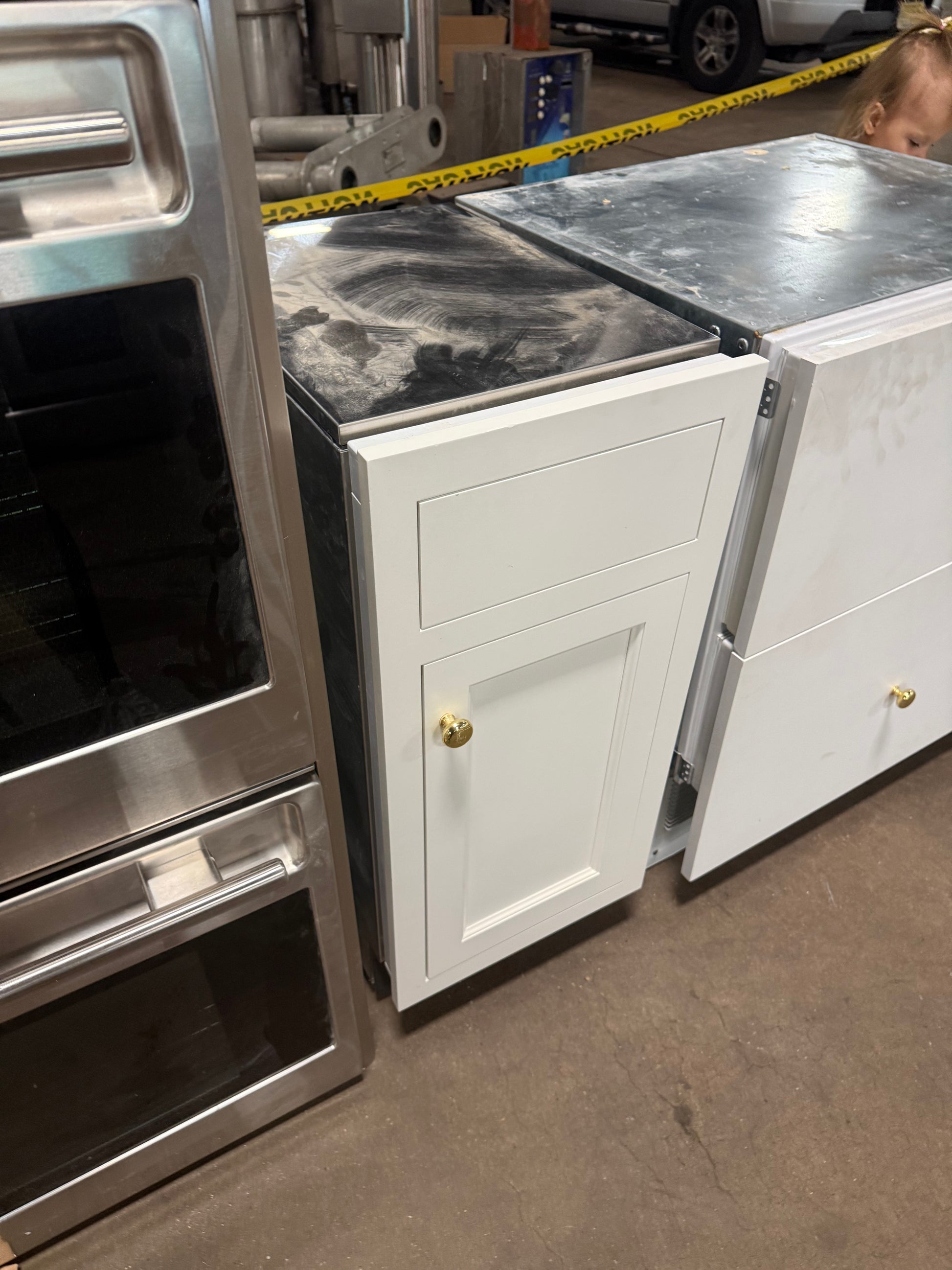 A white shaker-style undercounter cabinet with a single drawer and one lower door, both fitted with black countertops, shown in a warehouse or appliance showroom alongside stainless steel appliances. A person is partially visible in the upper right corner.