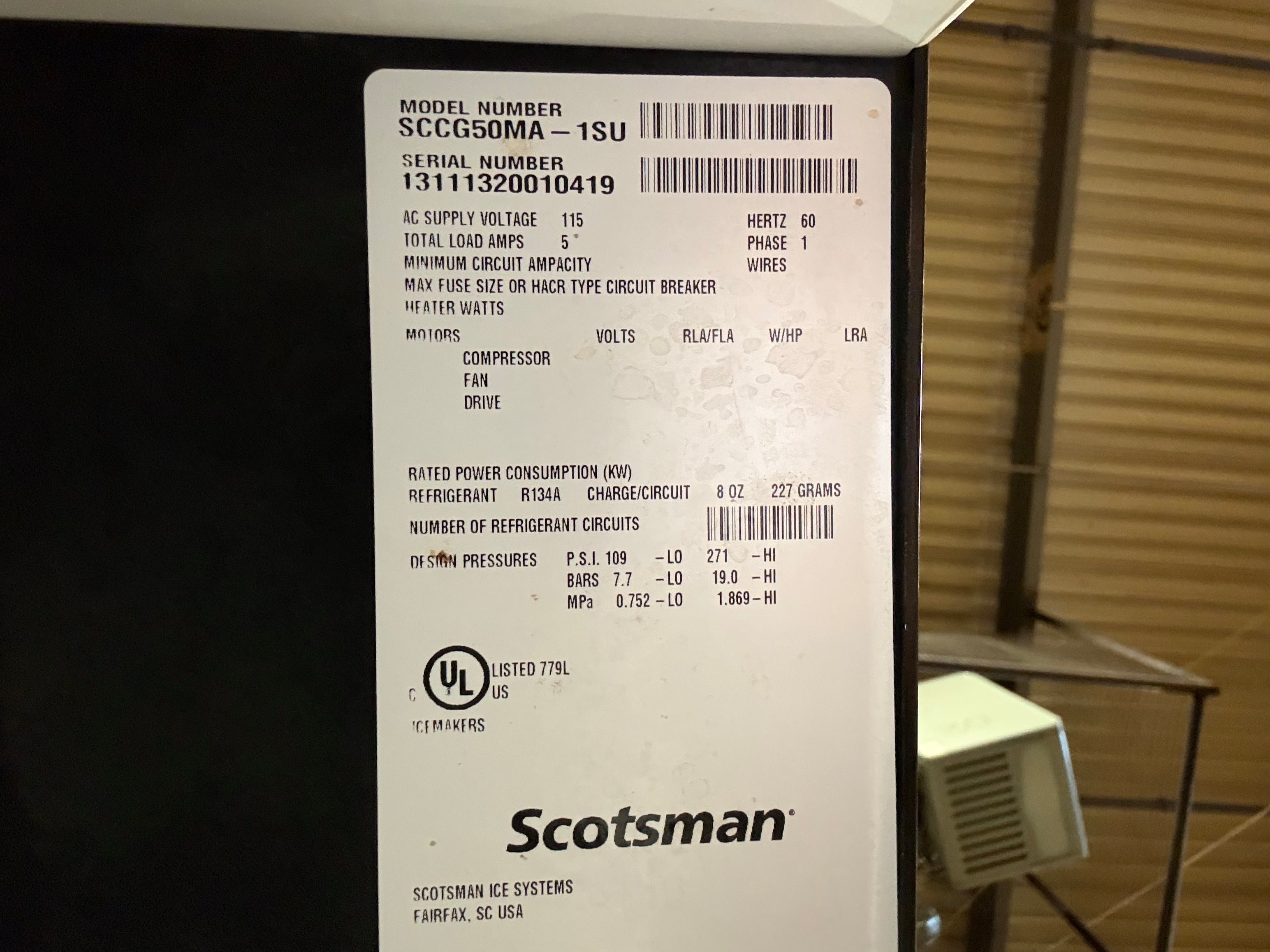 A close-up of a white Scotchman product label showing model information and specifications, attached to a black appliance in an indoor setting.