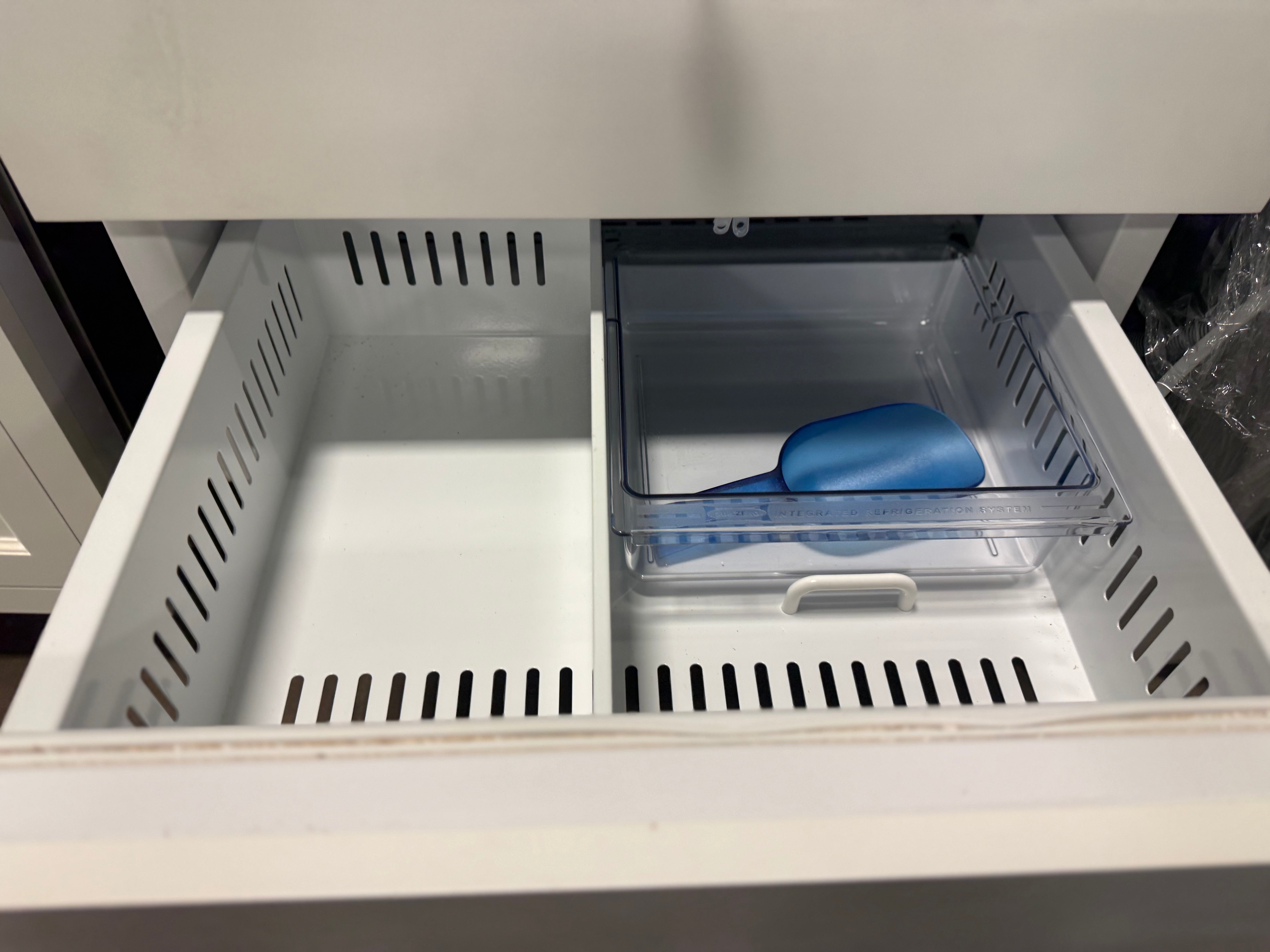 An open refrigerator drawer with two interior bins: a larger empty white bin on the left and a smaller clear bin on the right containing a blue scoop, set inside a white refrigerator.