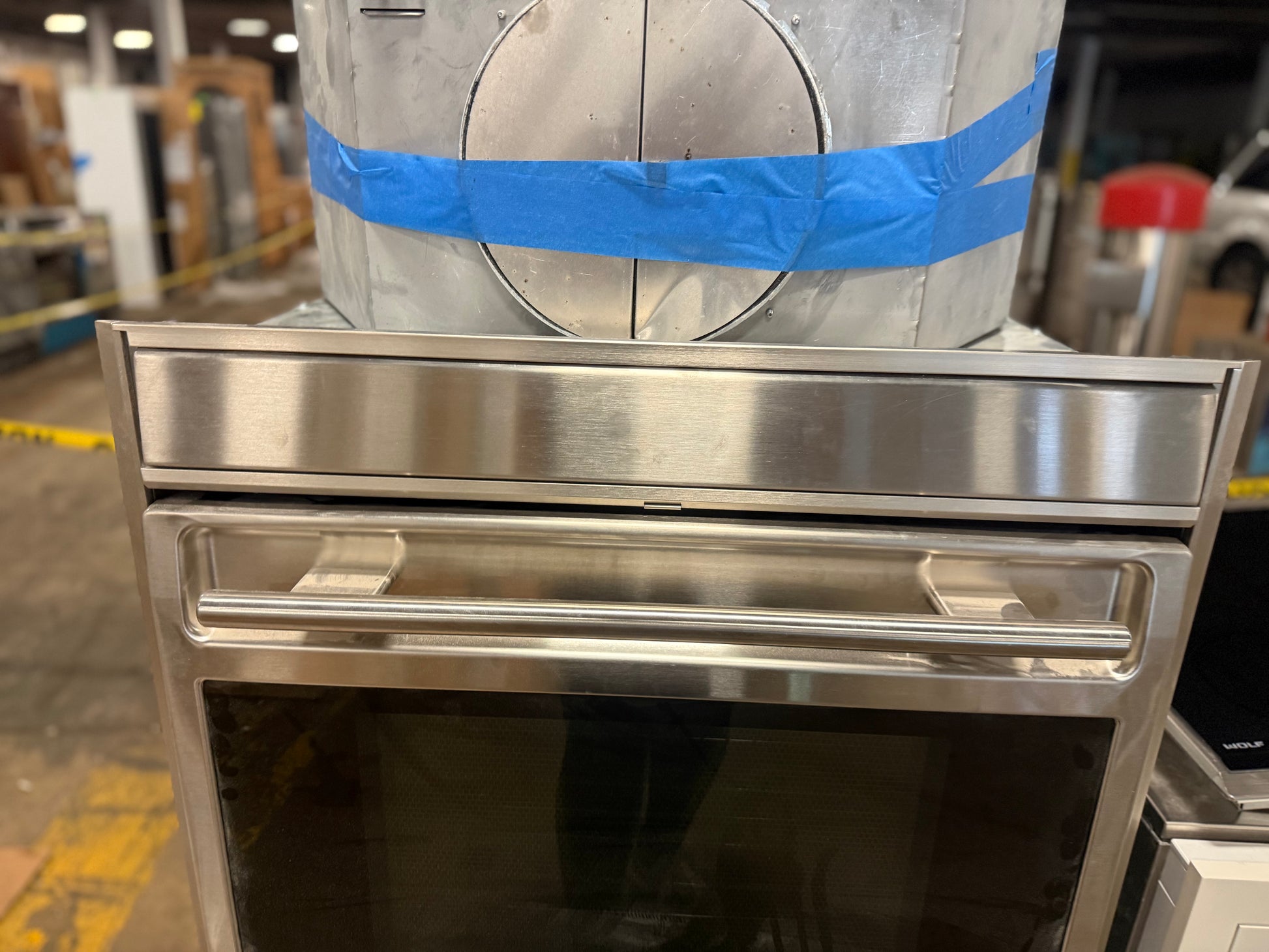 A stainless steel built-in oven or wall oven shown in a warehouse or showroom setting, with a large silver cylindrical duct wrapped in blue tape behind it.