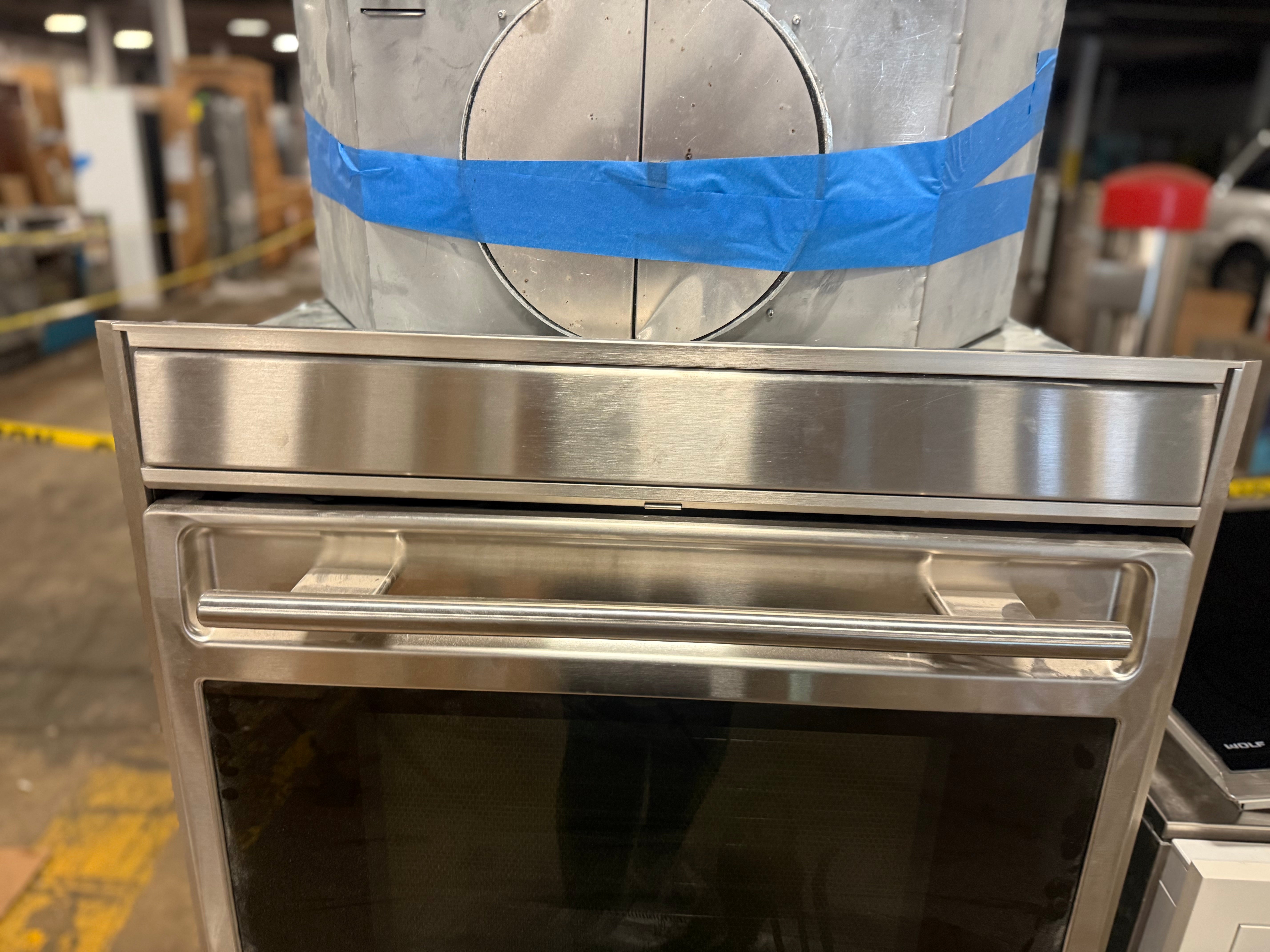 A stainless steel built-in oven or wall oven shown in a warehouse or showroom setting, with a large silver cylindrical duct wrapped in blue tape behind it.