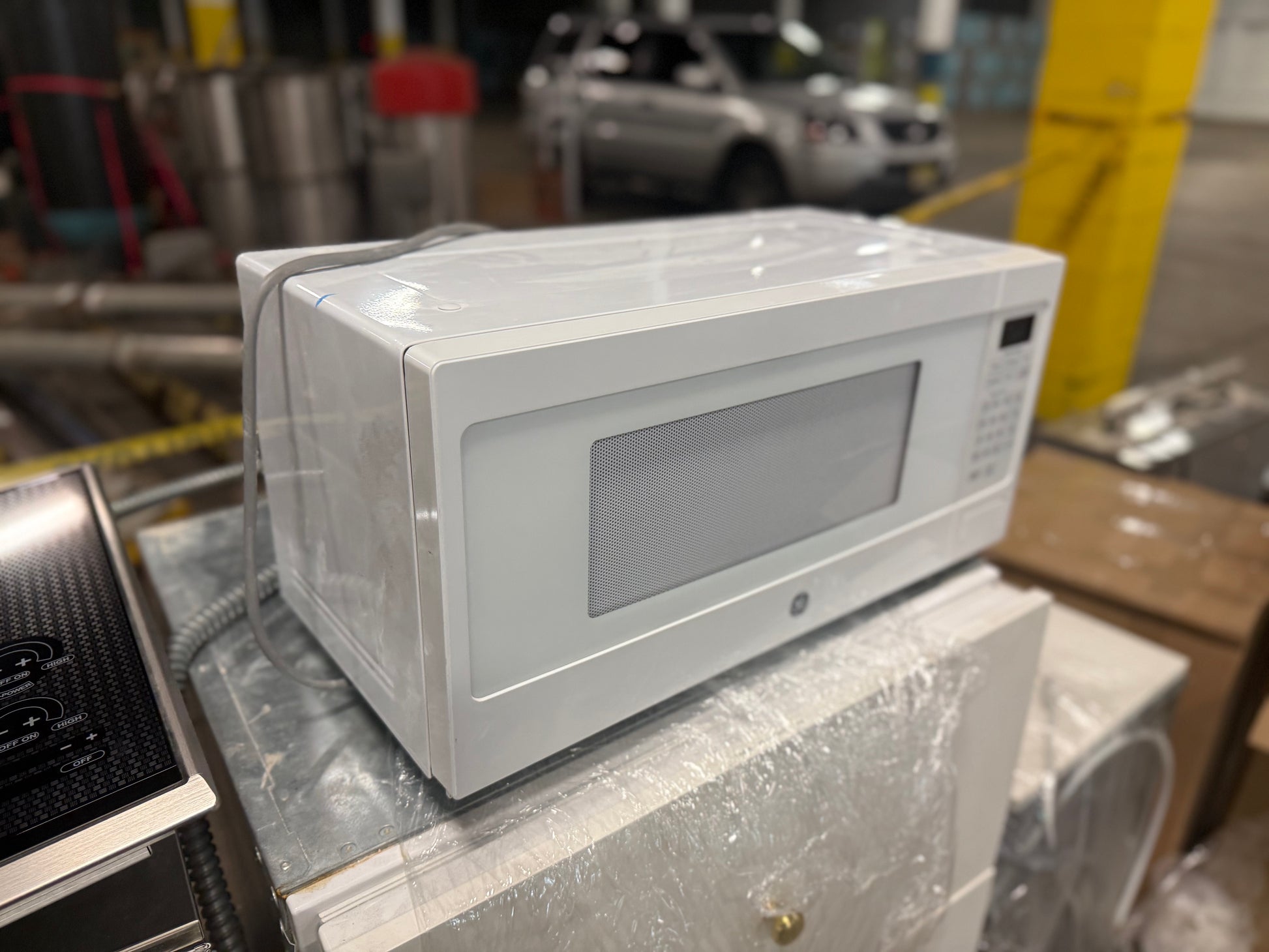 A white GE microwave oven wrapped in protective plastic, sitting on top of a wrapped appliance in what appears to be a warehouse or showroom setting.
