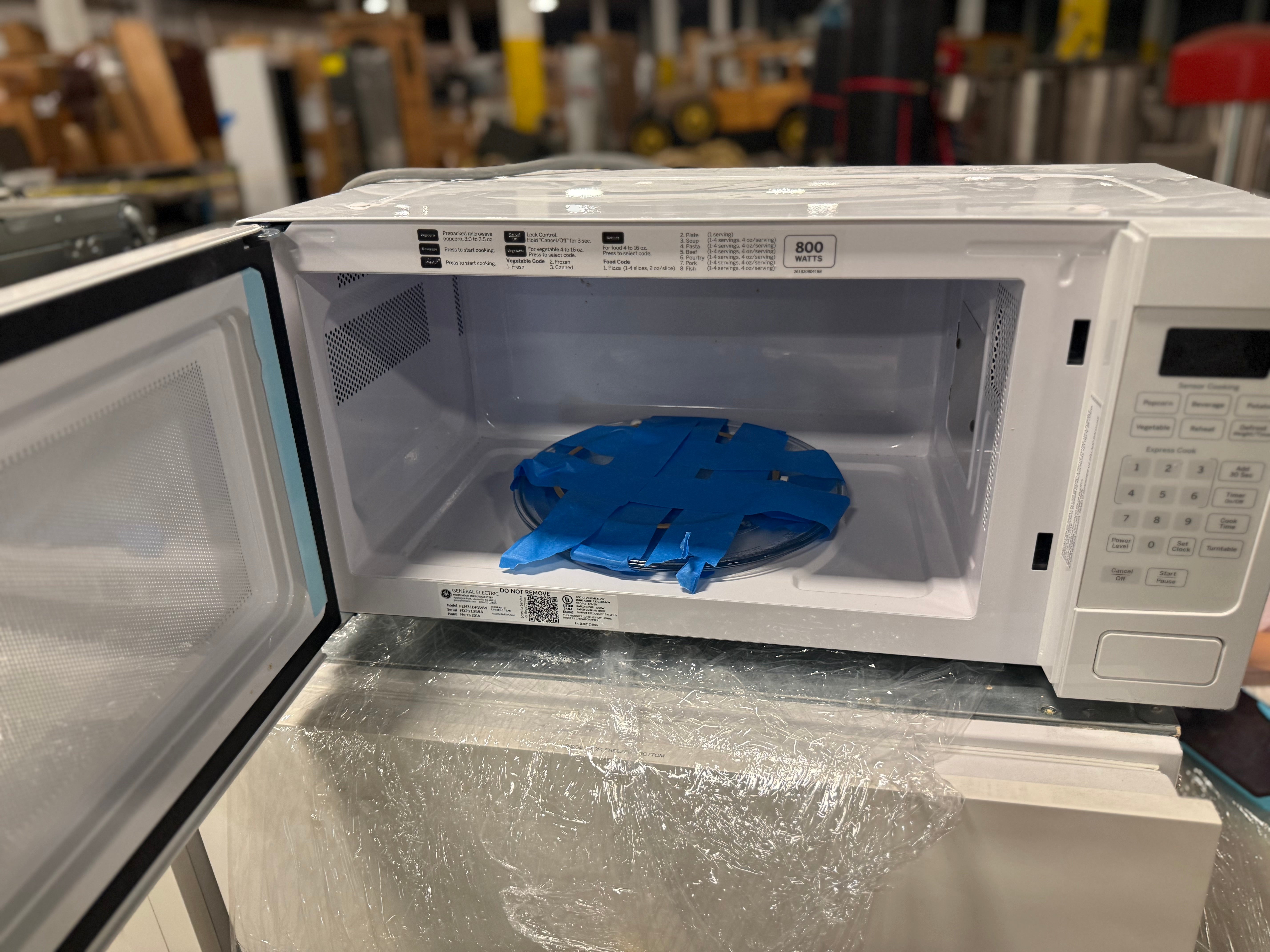 A white microwave oven with its door open, showing a blue silicone or plastic insert inside. The microwave is sitting on a reflective surface with a blurred workshop or store background.