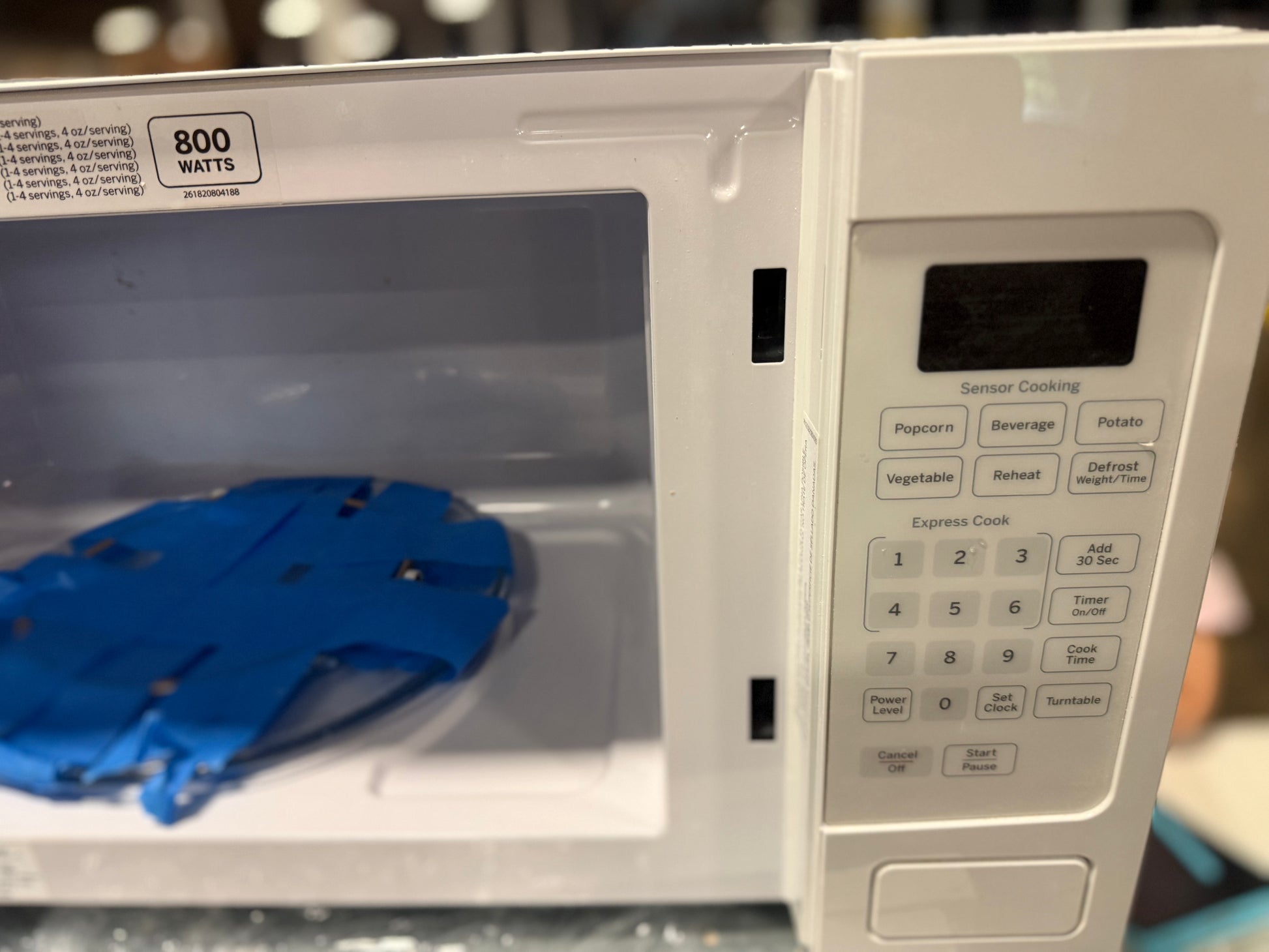 A white microwave oven with its door open, showing a blue plastic insert or tray inside. The control panel on the right has buttons for Popcorn, Beverage, Potato, Vegetable, Refresh, Defrost, and Express Cook, with an 800 watts rating sticker visible on the upper left.
