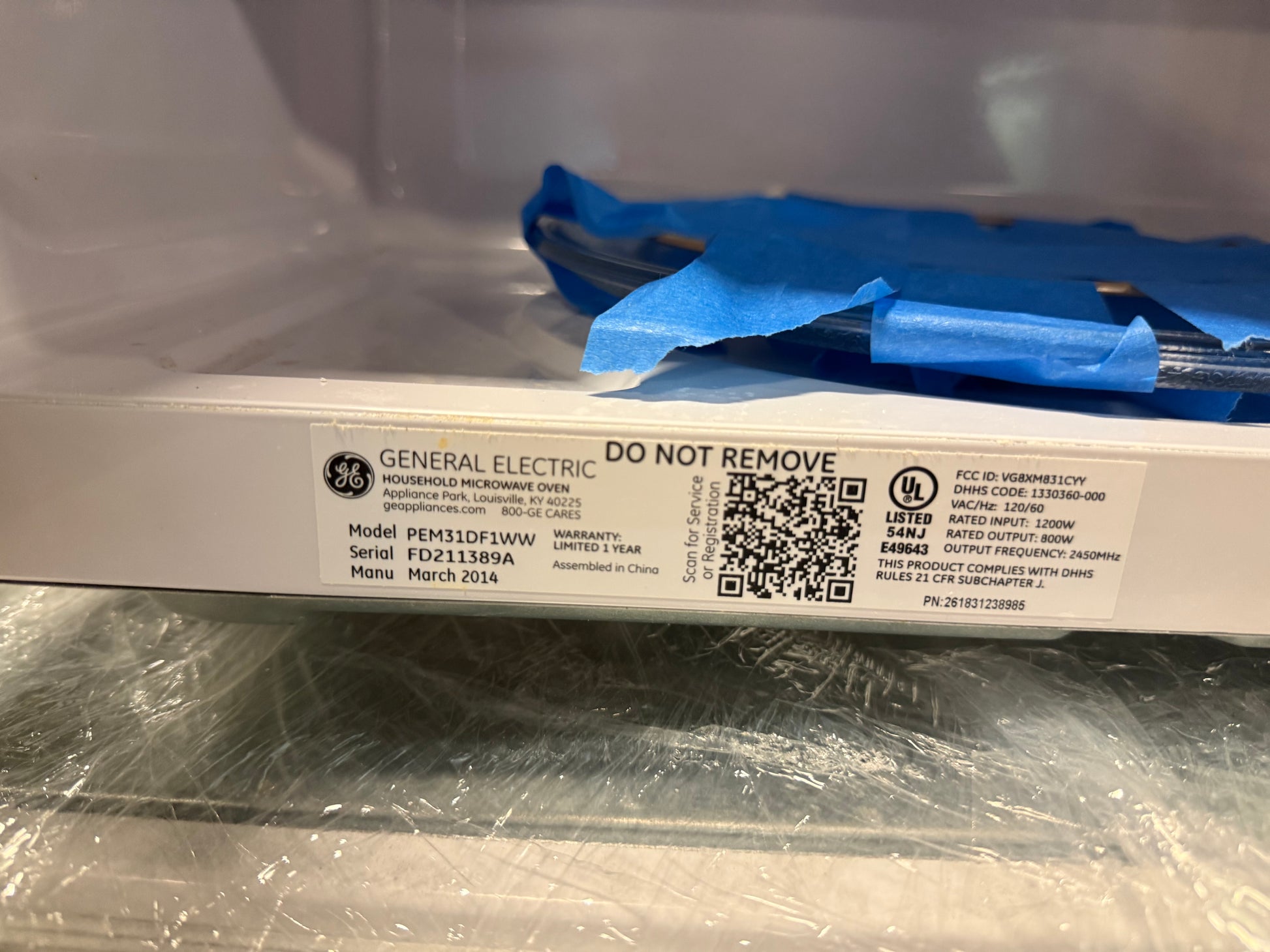 A close-up of a General Electric microwave door label showing model information, a QR code, and a blue protective covering on the inside glass door.