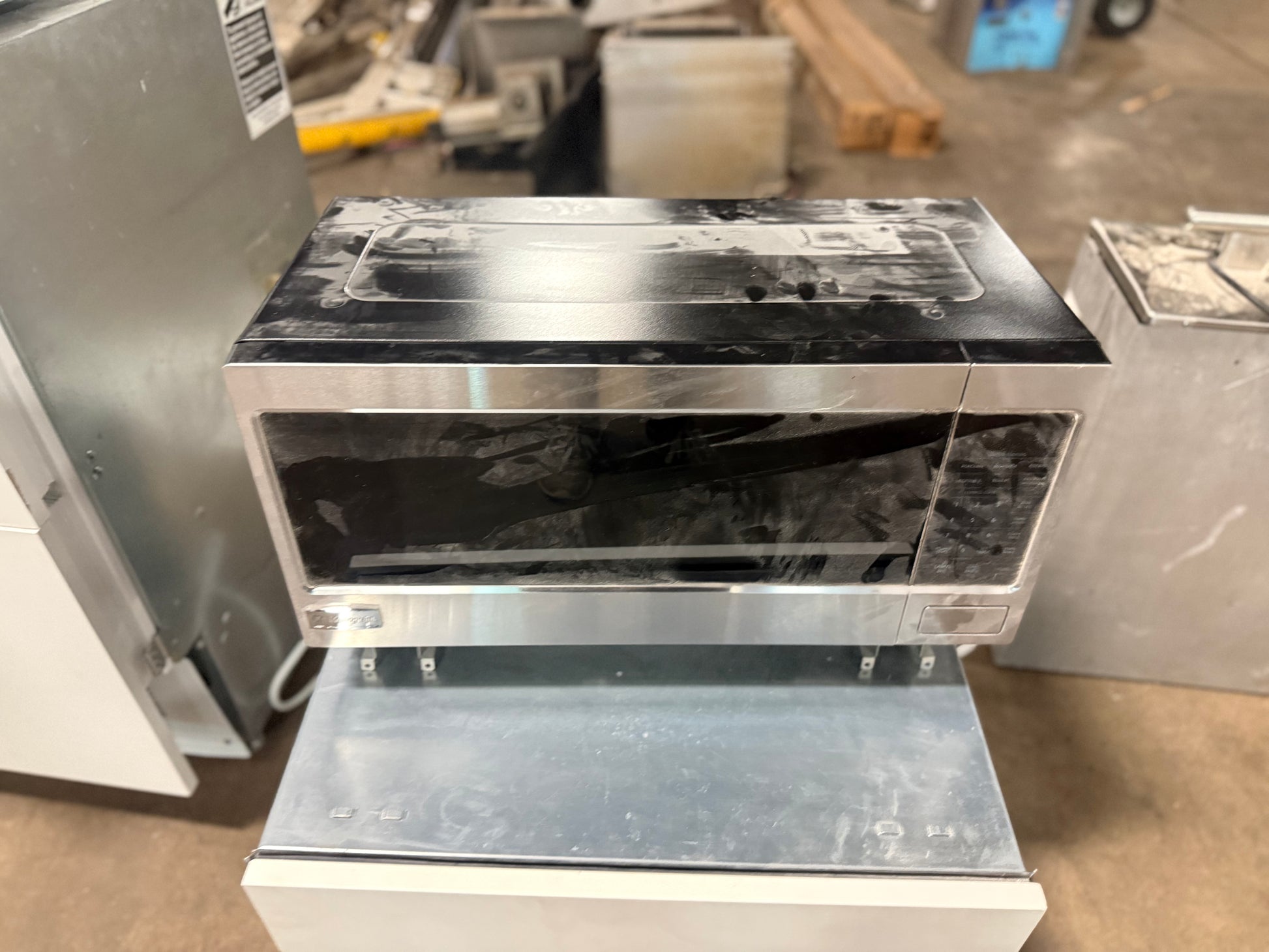 A stainless steel and black microwave oven with a protective plastic wrap, displayed on a metal support stand in what appears to be a warehouse or appliance repair shop.