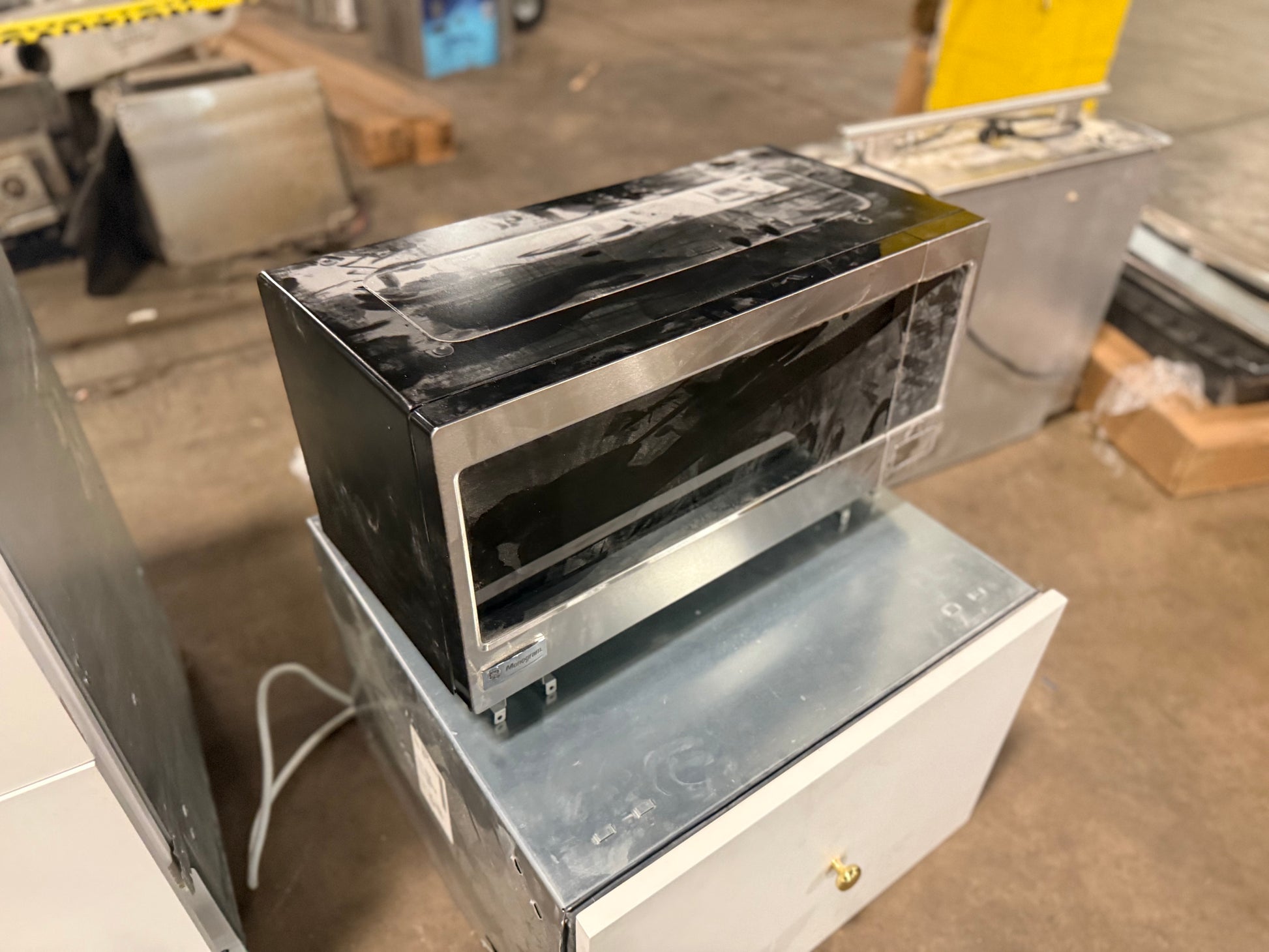 A black and stainless steel countertop microwave oven with protective plastic still on top, sitting on a galvanized metal cabinet in what appears to be a warehouse or appliance showroom.