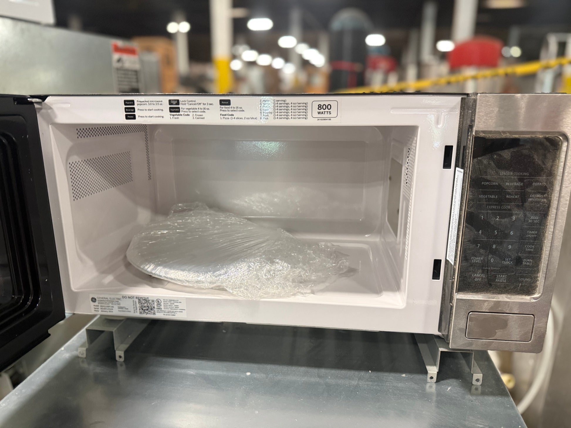 A white and stainless steel microwave oven with its door open, showing the interior door panel, glass window, and control panel. A plastic-wrapped item sits inside the microwave on a metal surface in what appears to be a warehouse or showroom setting.