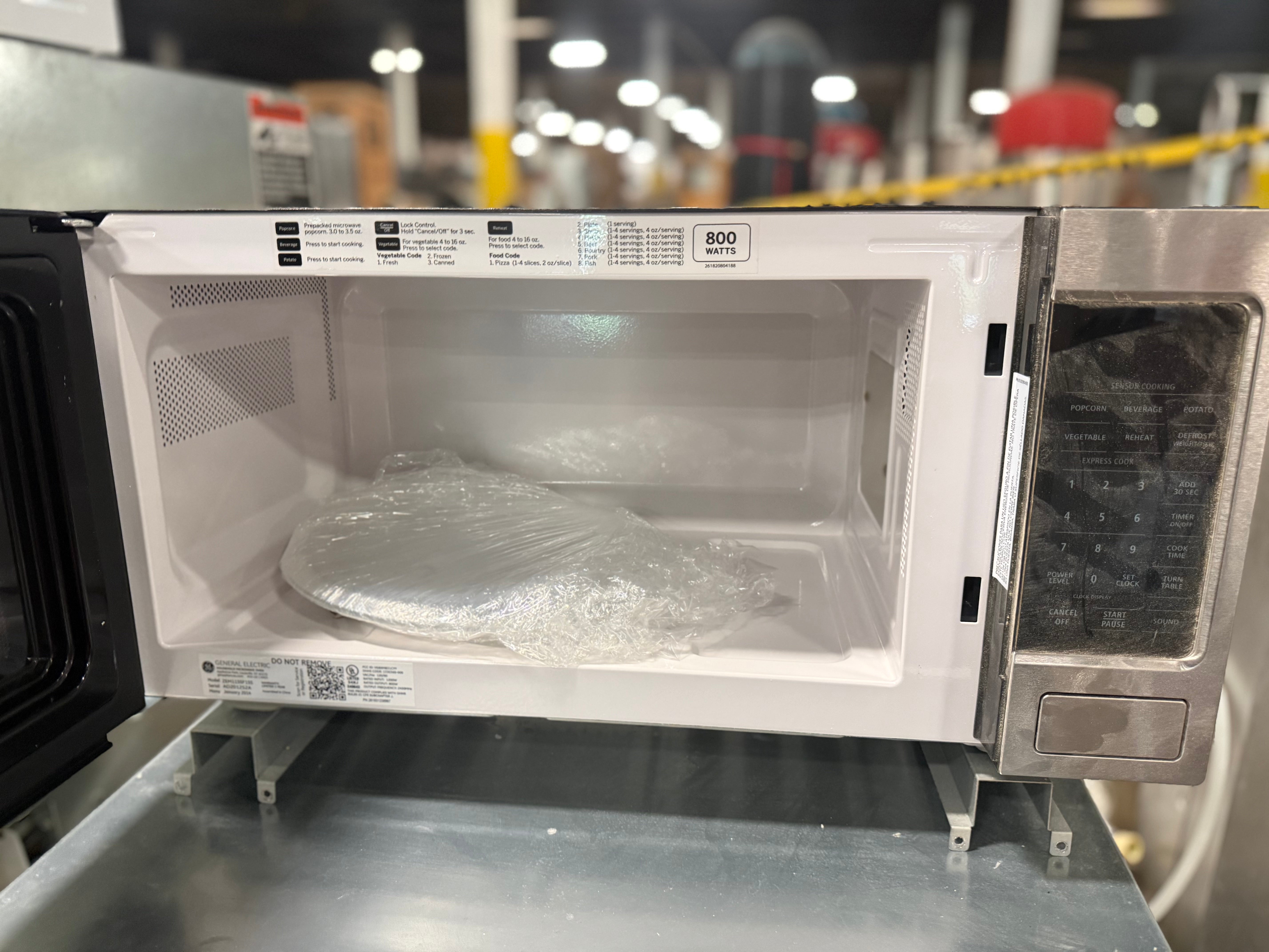 A white and stainless steel microwave oven with its door open, showing the interior door panel, glass window, and control panel. A plastic-wrapped item sits inside the microwave on a metal surface in what appears to be a warehouse or showroom setting.