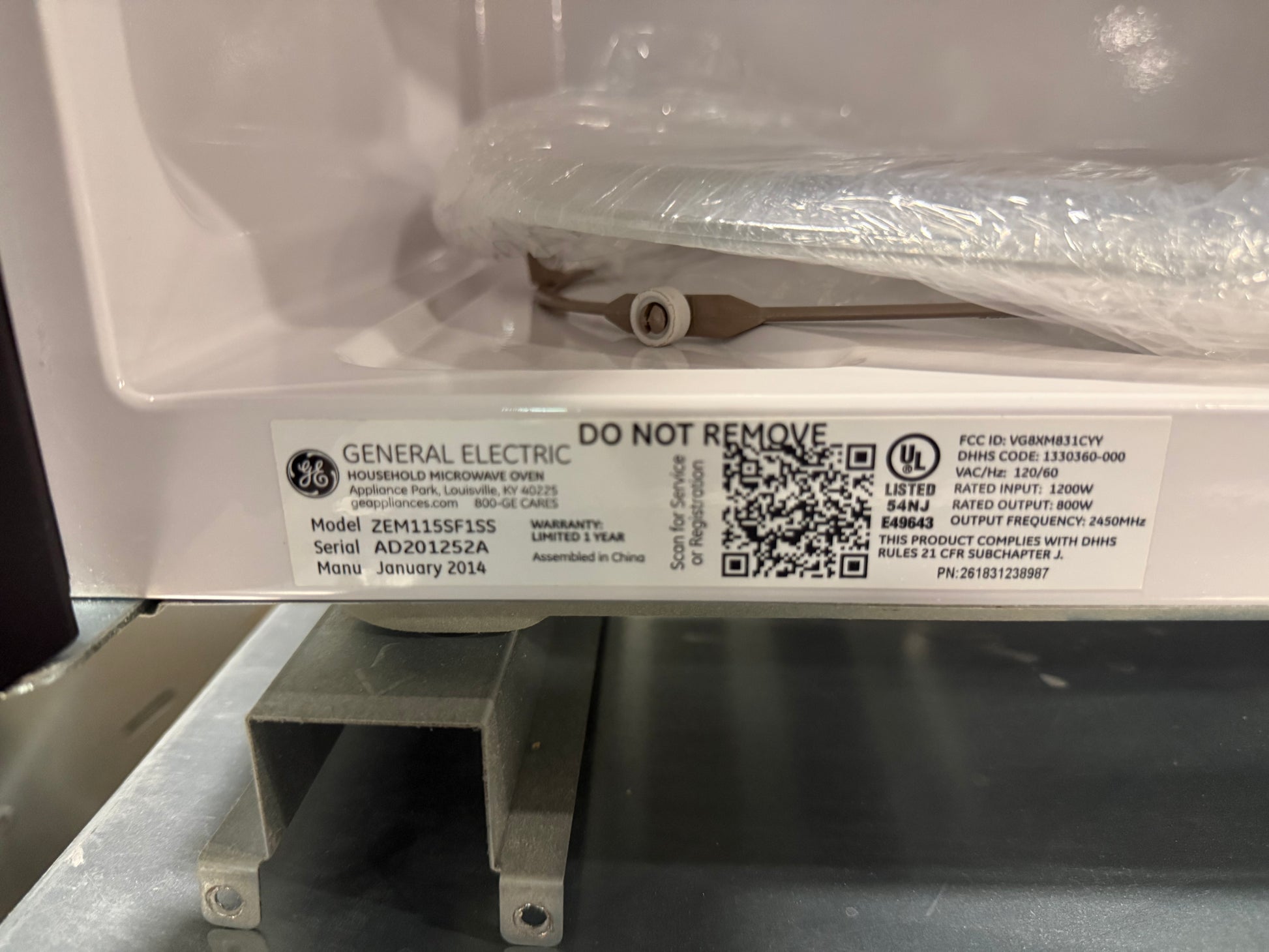 A close-up of a General Electric water heater label on the white metal front panel, with a clear protective plastic wrap still covering part of the upper unit. The label shows model information, a QR code, and compliance markings.