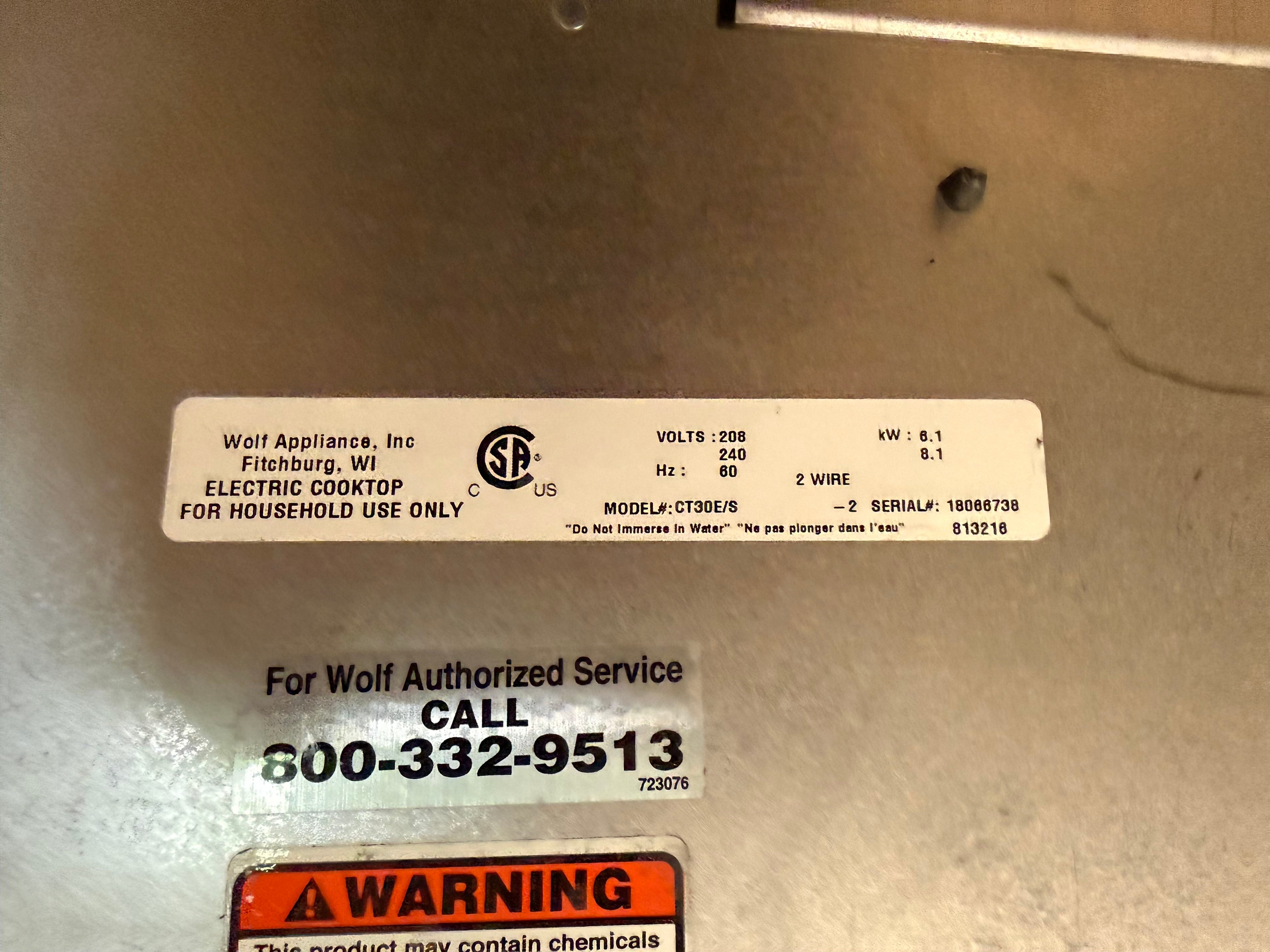 A close-up of a stainless steel appliance panel with a white product information label and a warning sticker. The label reads Wolf Appliance, Inc and shows model details for an electric cooktop.