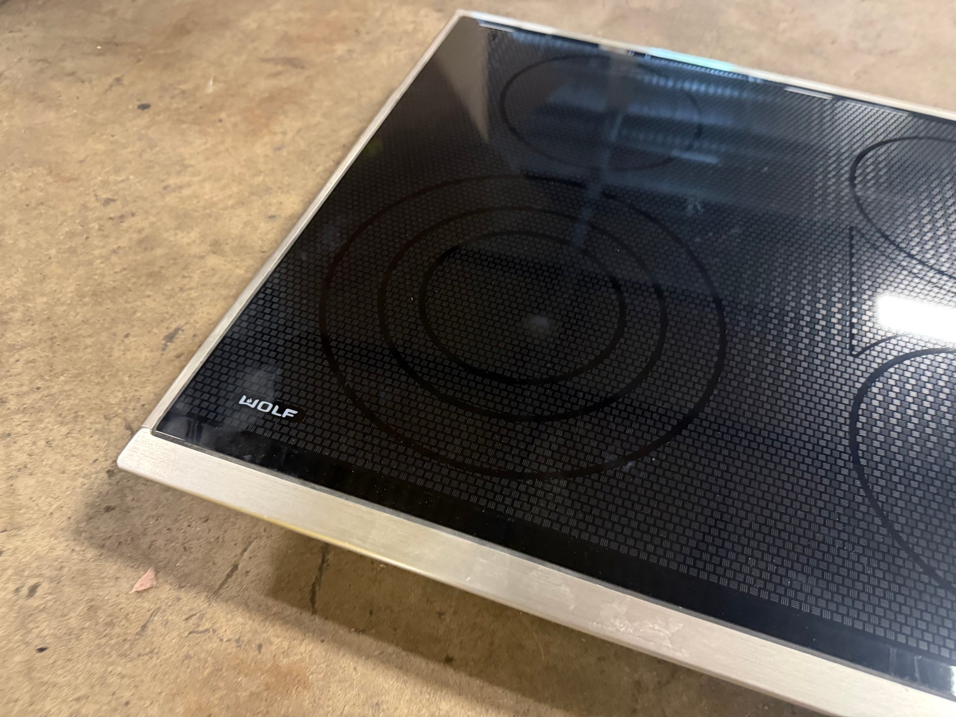 A Wolf brand glass-ceramic electric cooktop with a black induction surface and silver metal frame, photographed on a concrete floor.