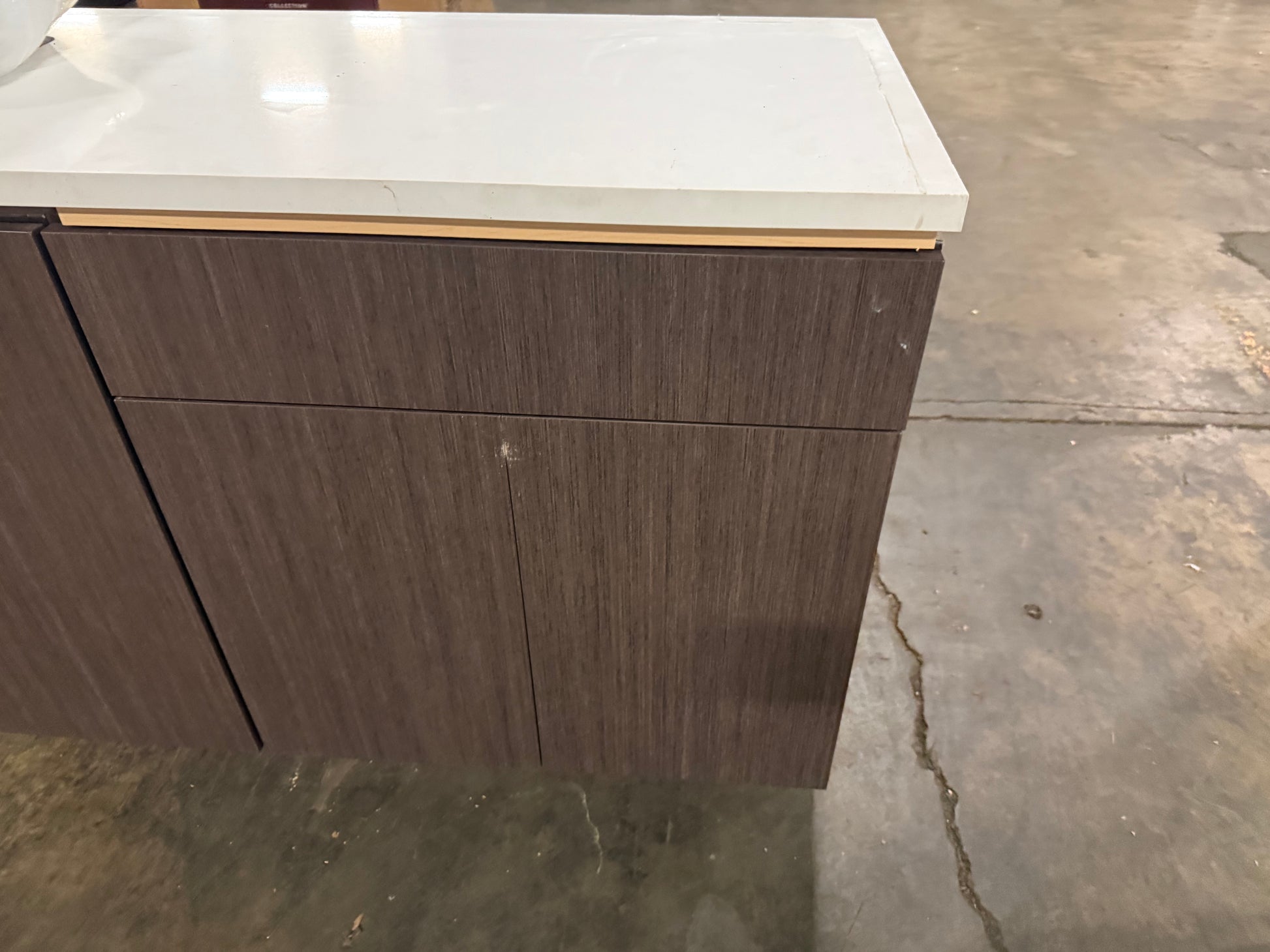 A floating kitchen cabinet with dark brown wood-grain doors and a white countertop, shown in a warehouse or showroom setting on a concrete floor.