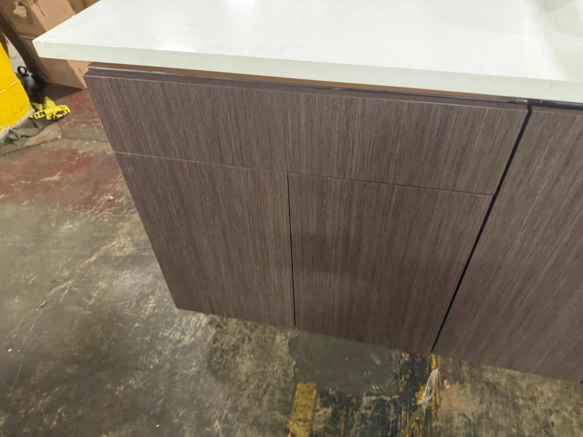 A modern kitchen cabinet with dark brown wood grain doors and a white countertop, shown in a warehouse or workshop setting with a concrete floor.