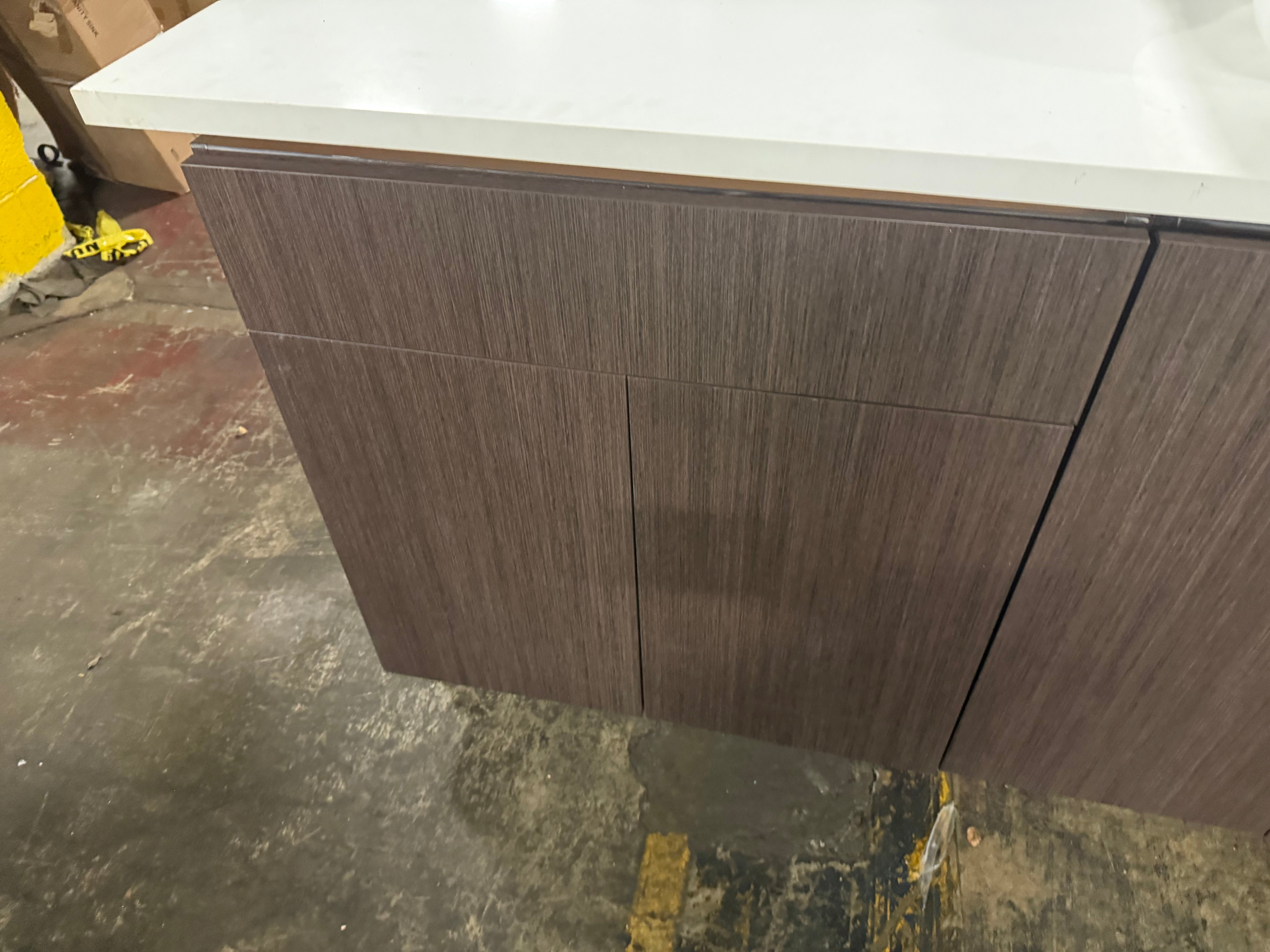 A modern kitchen cabinet with dark brown wood grain doors and a white countertop, shown in a warehouse or workshop setting with a concrete floor.
