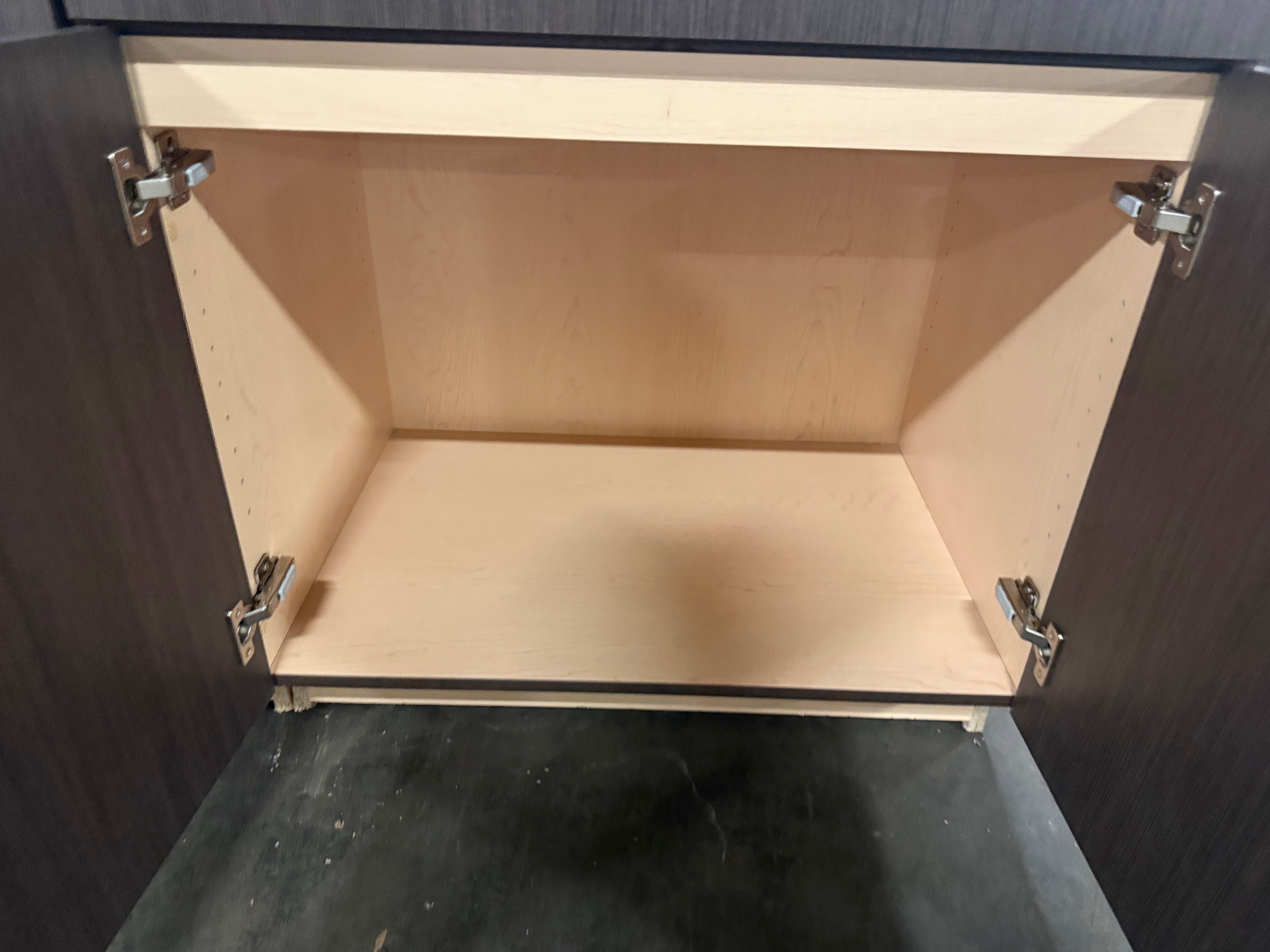 An open dark wood kitchen cabinet showing a light natural wood interior shelf and silver metal hinges.
