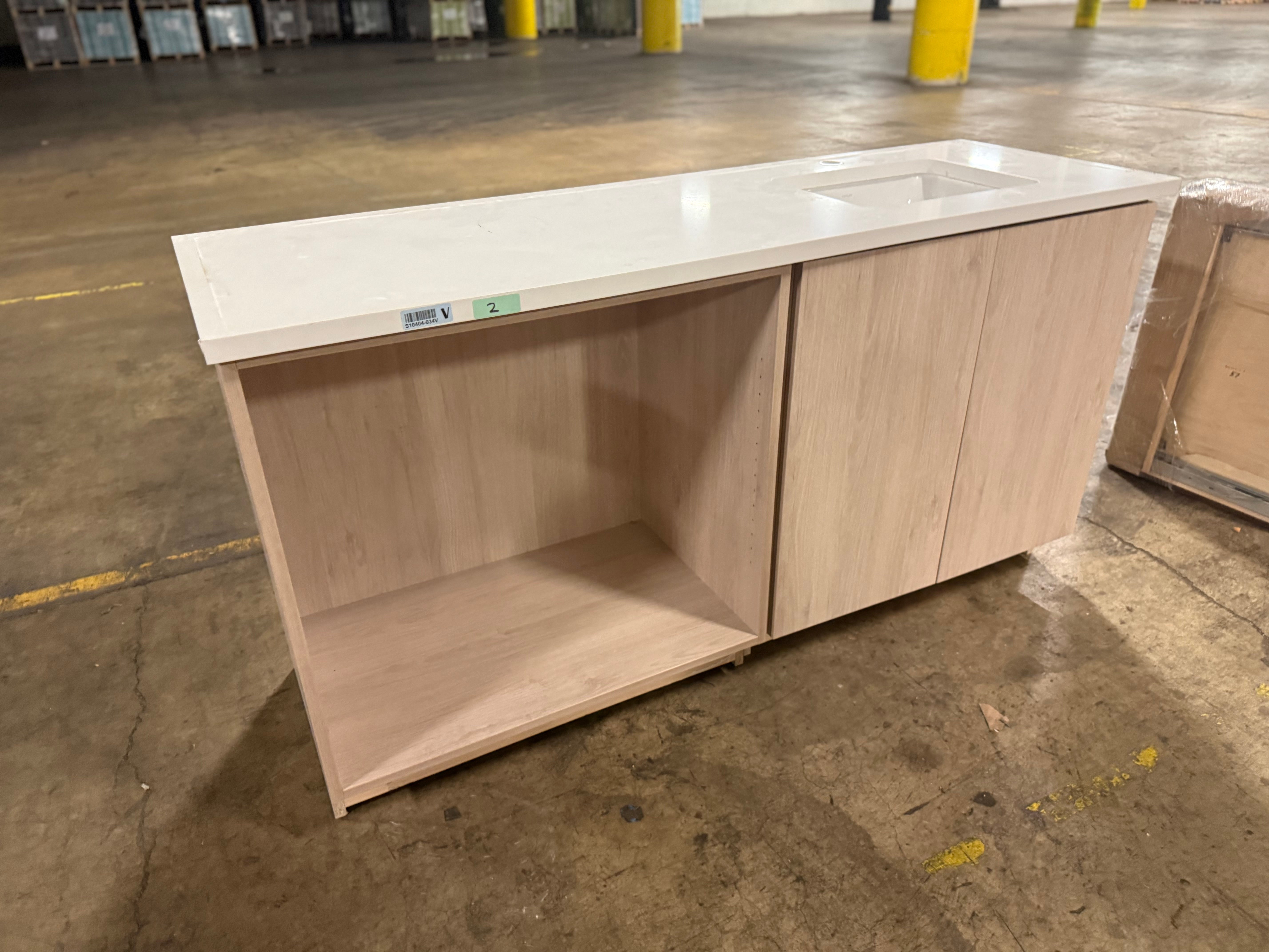 A light wood kitchen island cabinet with a white countertop and integrated sink, photographed in a warehouse setting on a concrete floor.